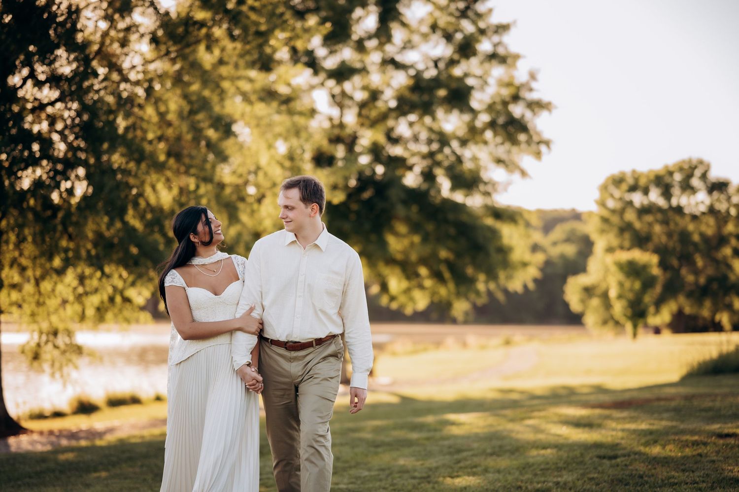 Couple in elegant attire walking hand-in-hand on lush green lawn with golden sunlight filtering through trees.