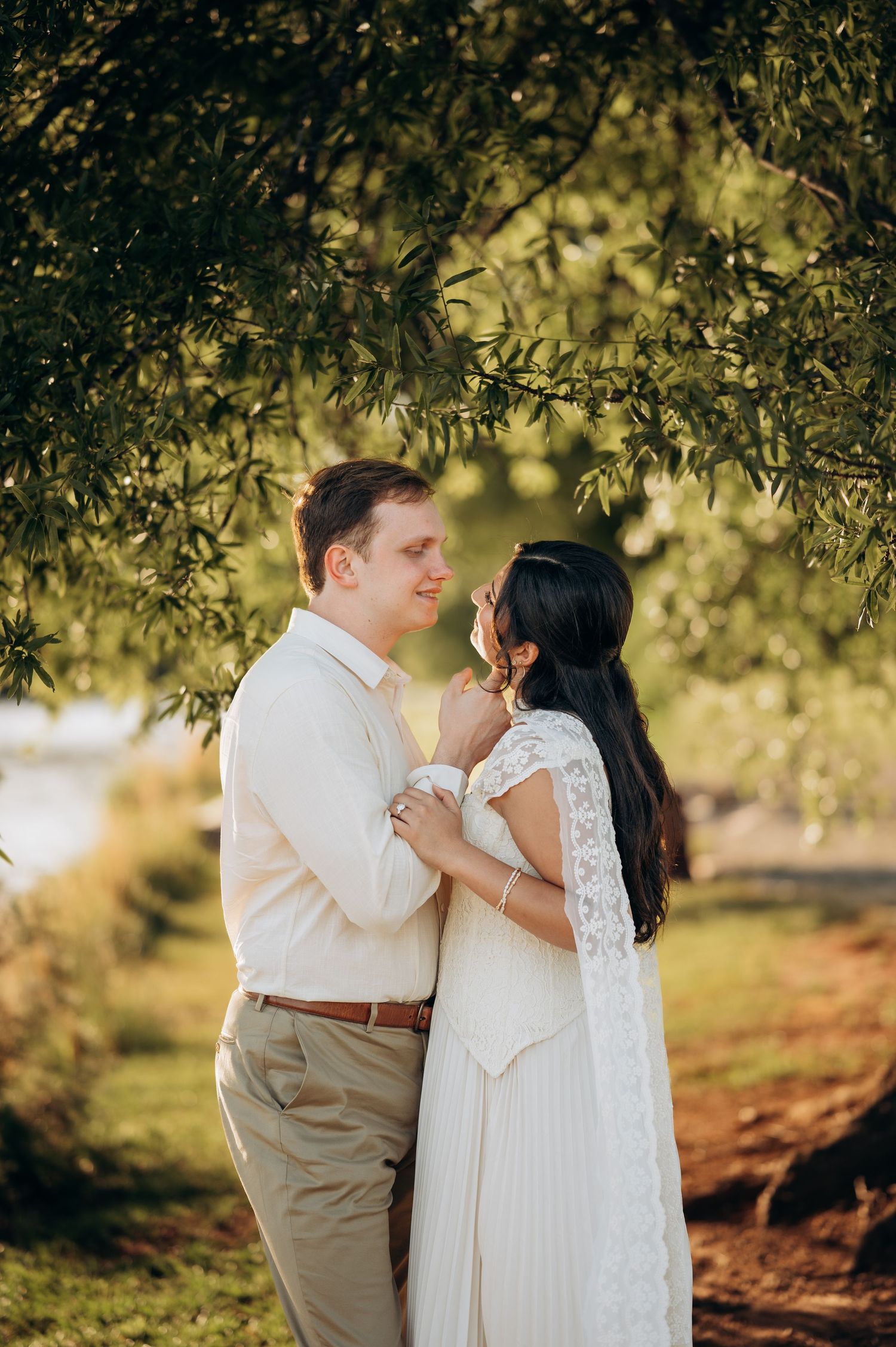 Couple in white and beige attire share a romantic moment under lush green tree branches in outdoor setting.