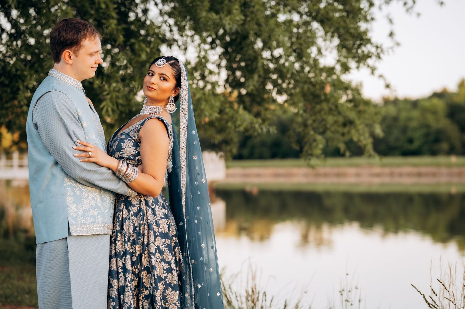 Couple in Indian wedding attire poses by a lake, woman in floral lehenga with blue dupatta, man in light blue sherwani.