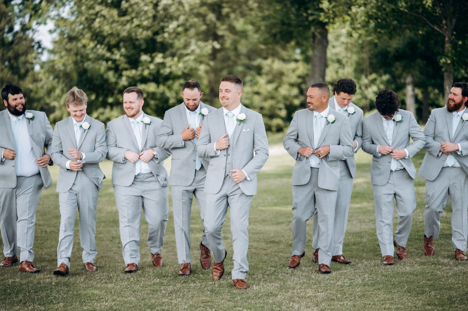 Groomsmen in matching light gray suits with boutonnieres walk outdoors on green grass at a wedding celebration.