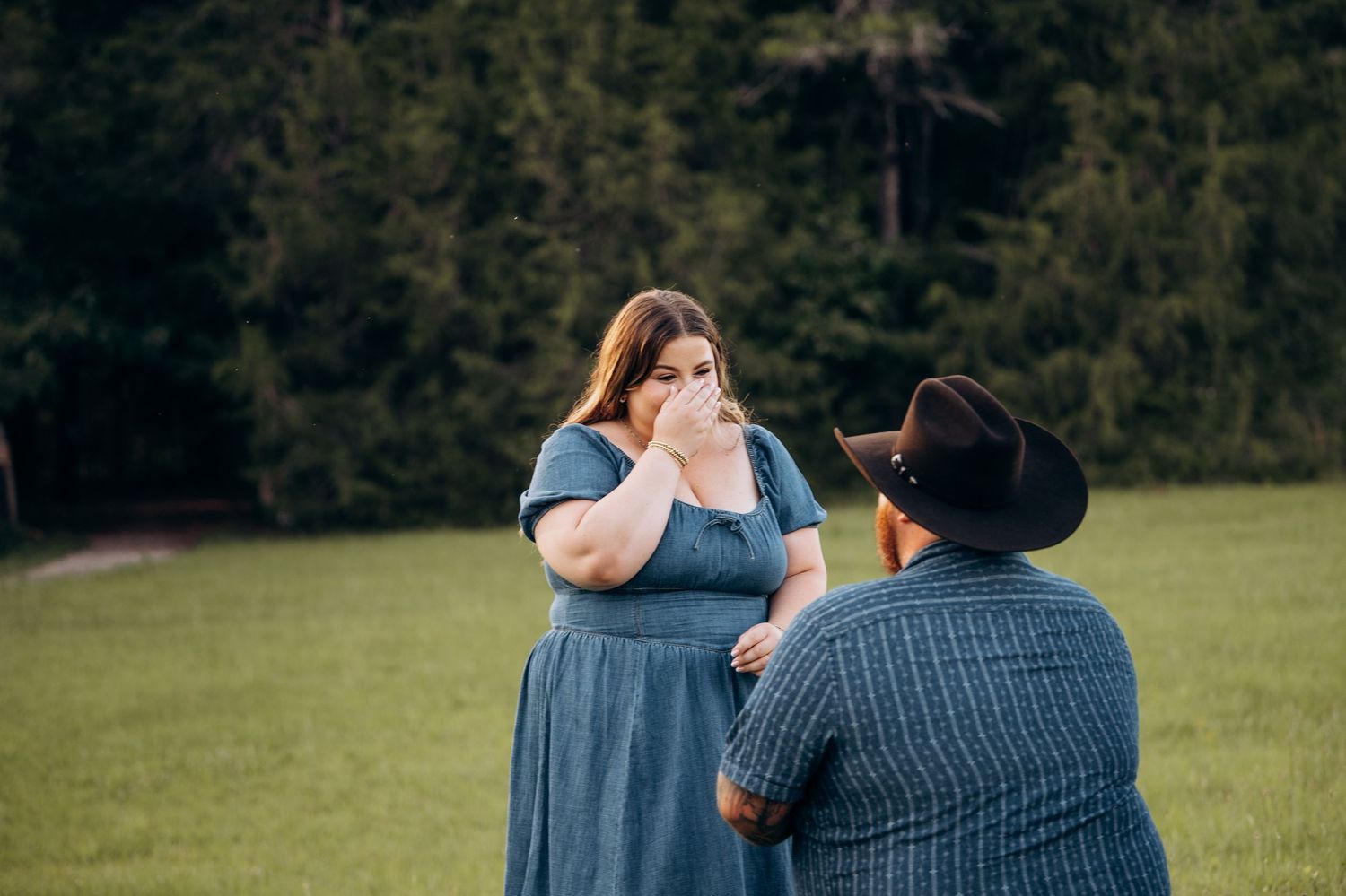 Emotional woman in blue dress covers her face while man in cowboy hat holds her hands in a field.