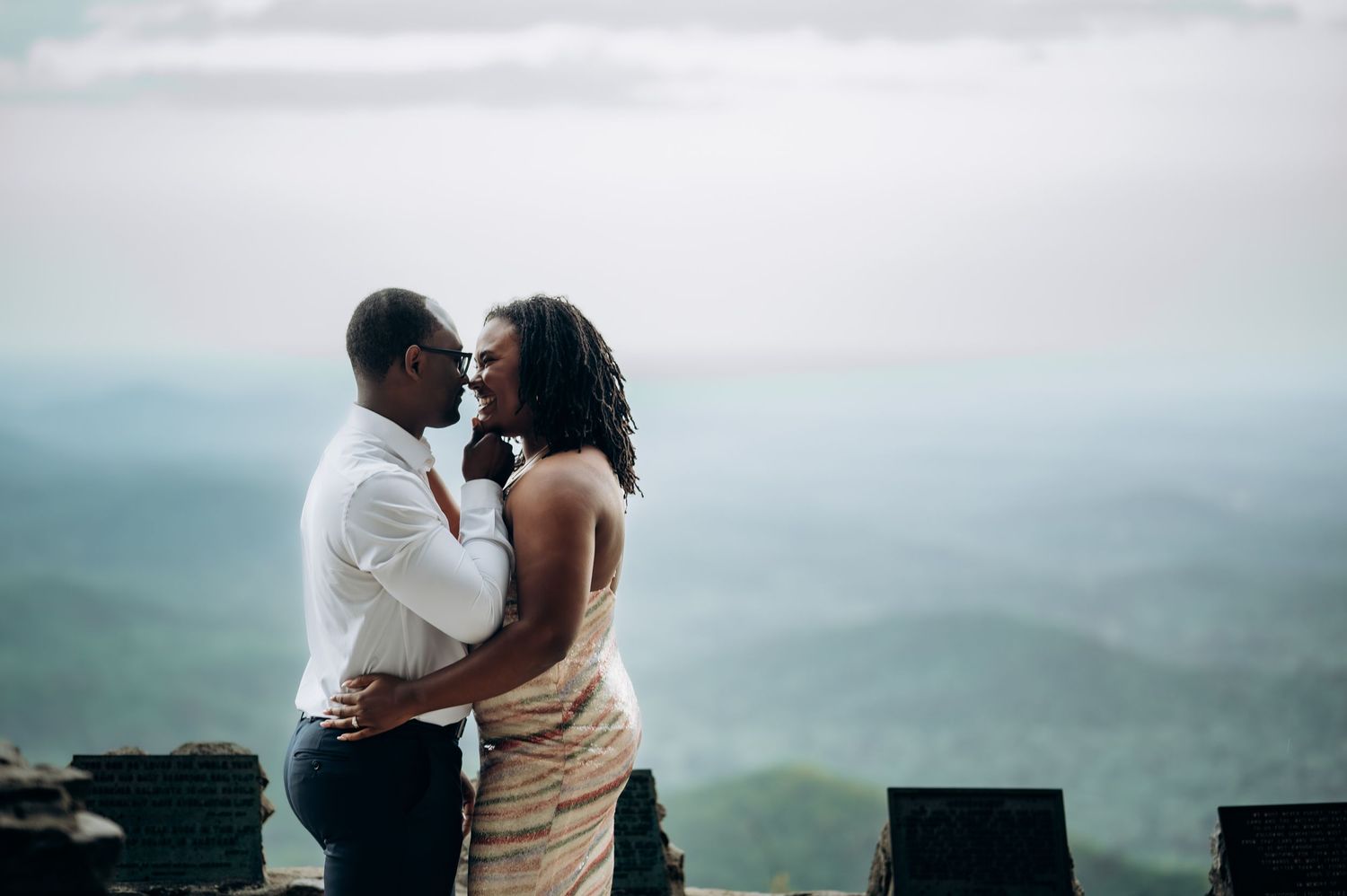 Couple sharing a romantic embrace on a scenic overlook with misty mountain views in the background.