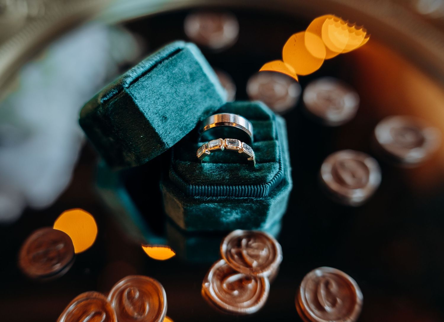 Two gold wedding rings displayed in a teal velvet ring box, surrounded by decorative wax seals with bokeh lighting.