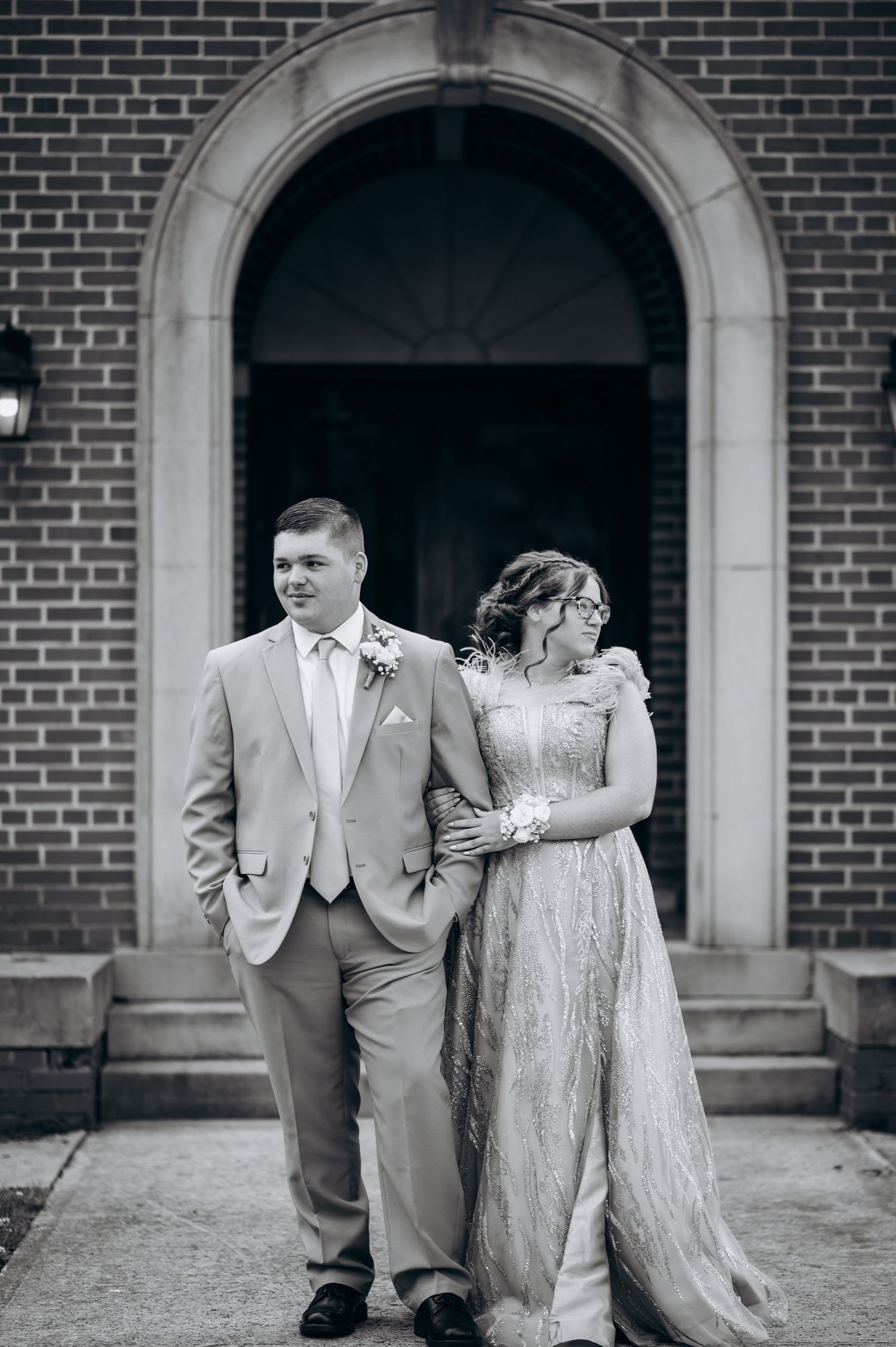 Black and white photo of a couple in formal attire posing before a brick church arch, woman in gown, man in suit.