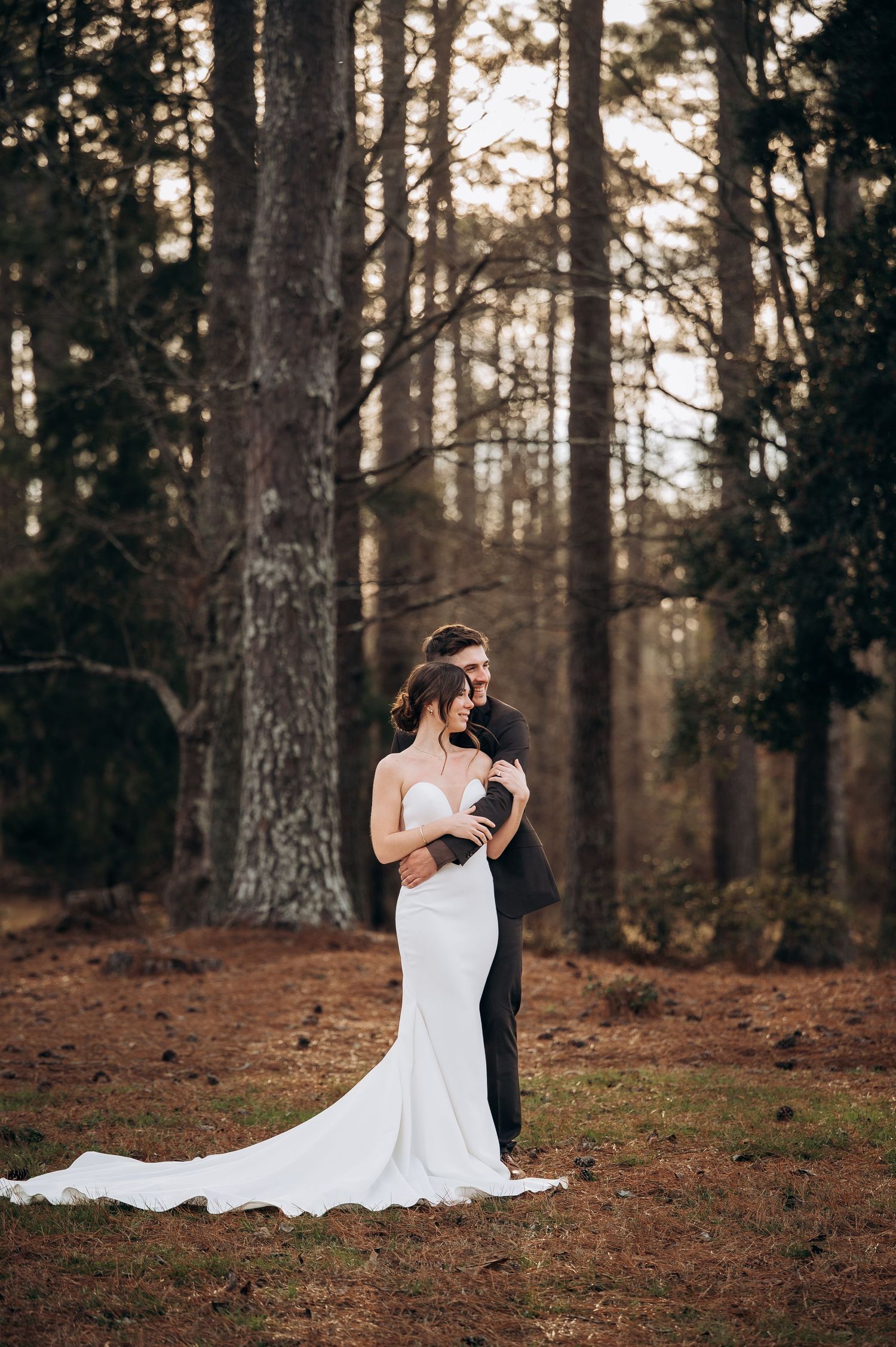 Bride and groom embrace romantically in a pine forest, bride wearing elegant white mermaid gown with long train.