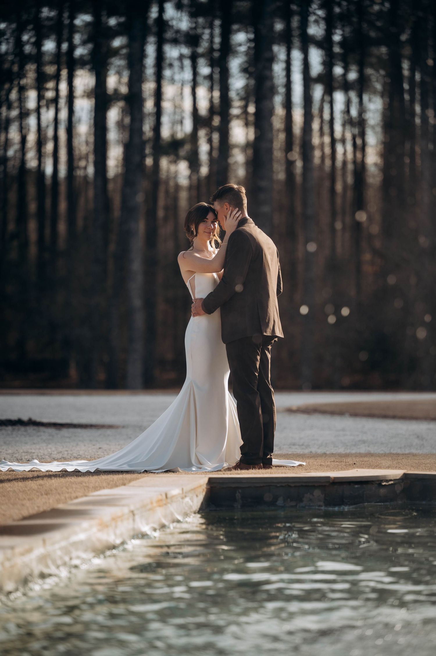 Bride and groom share a romantic moment by a reflective pool surrounded by tall trees in golden hour light.