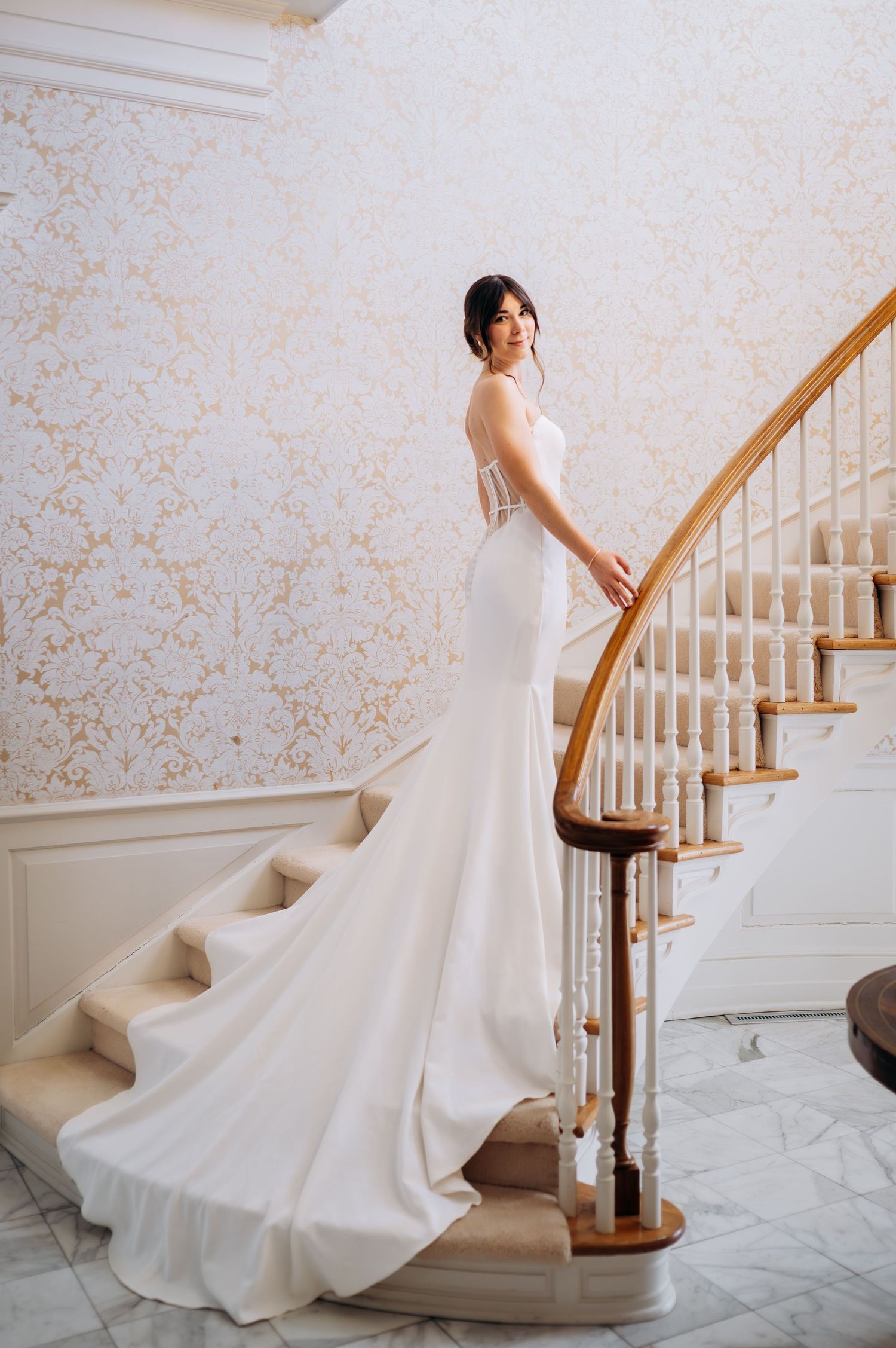 Stunning bride in backless white mermaid gown with long train posed on elegant curved staircase.