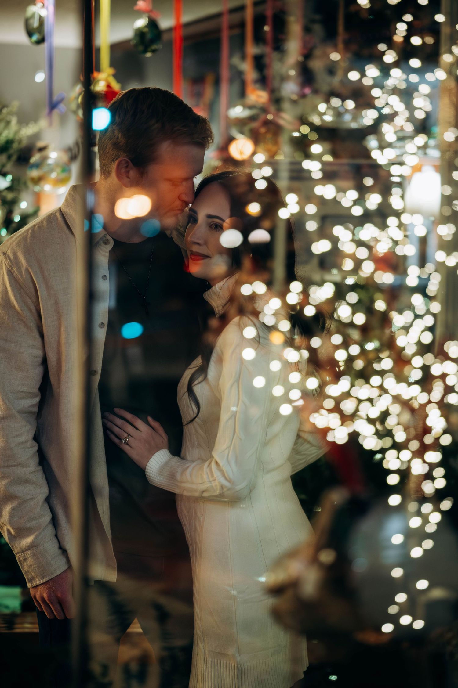 Romantic couple embracing and smiling near a glowing bokeh Christmas tree during a holiday pregnancy announcement.