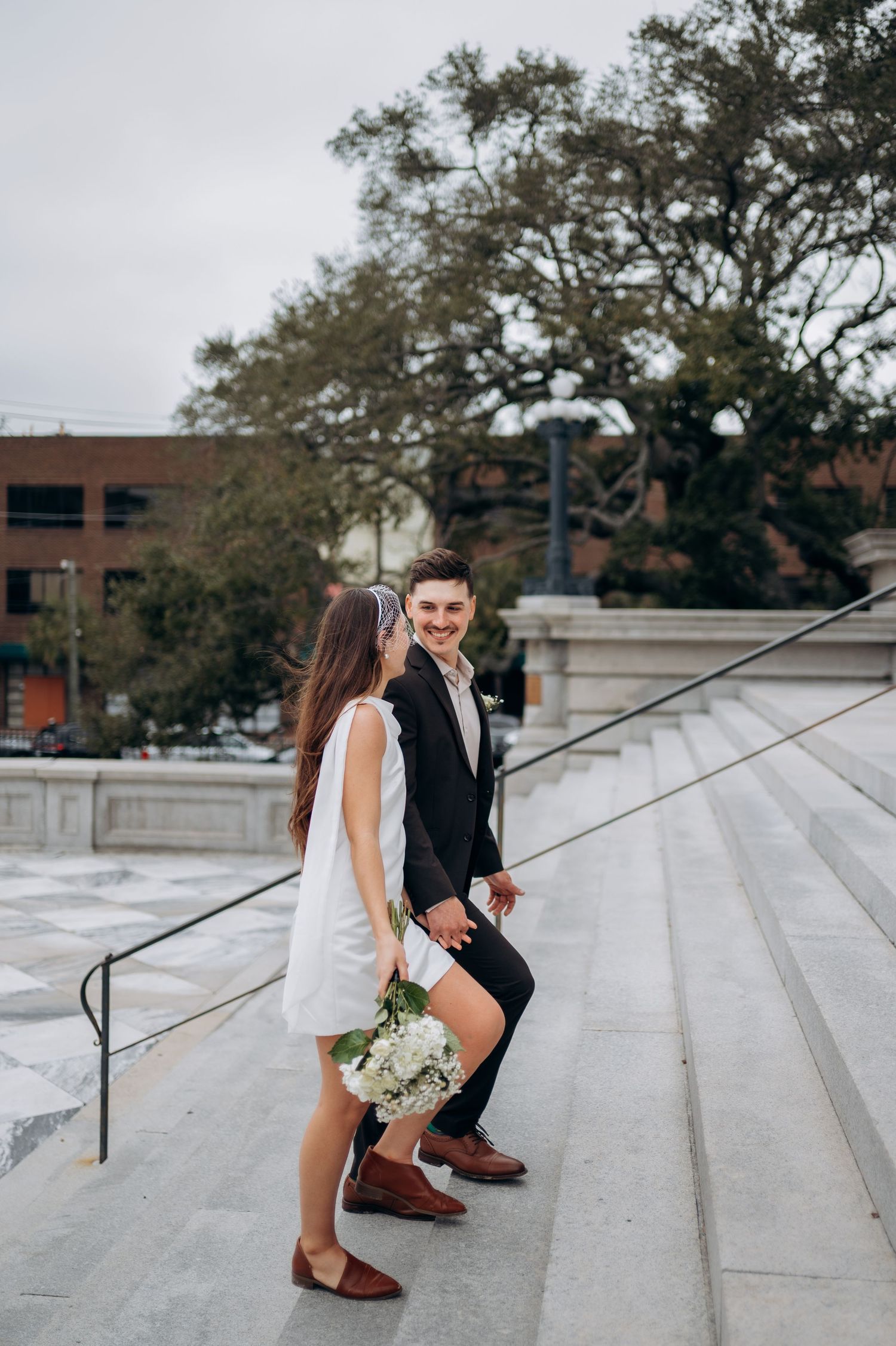 Couple in wedding attire walking up marble steps, bride holding white floral bouquet, groom smiling back at camera.