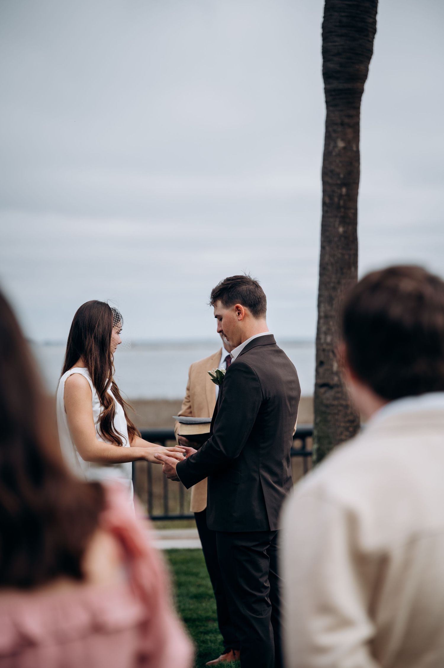 Couple exchanging vows at an outdoor wedding ceremony near the ocean, with guests watching nearby.