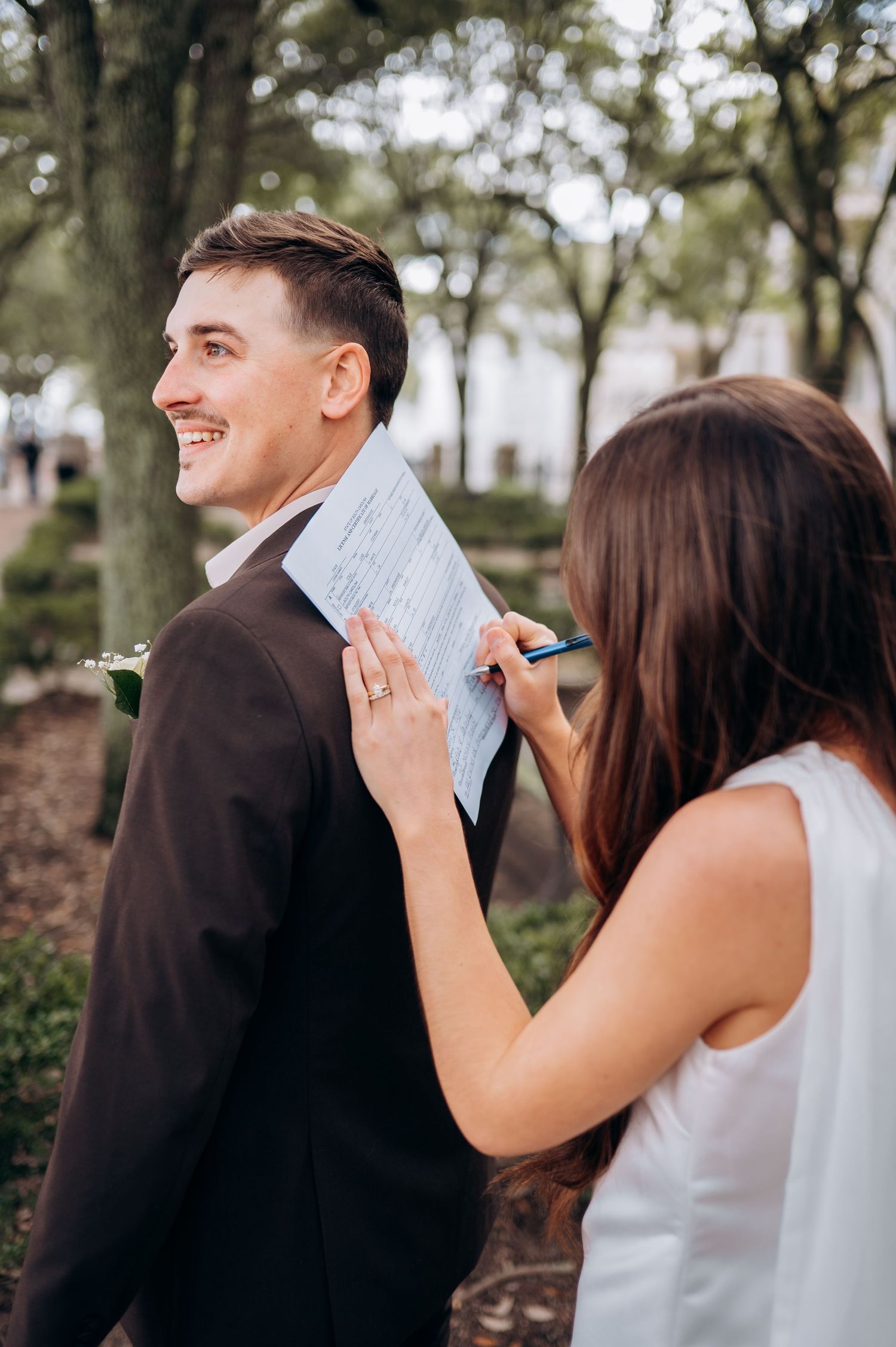 Smiling groom in dark suit watches bride sign marriage certificate using his back as a writing surface.