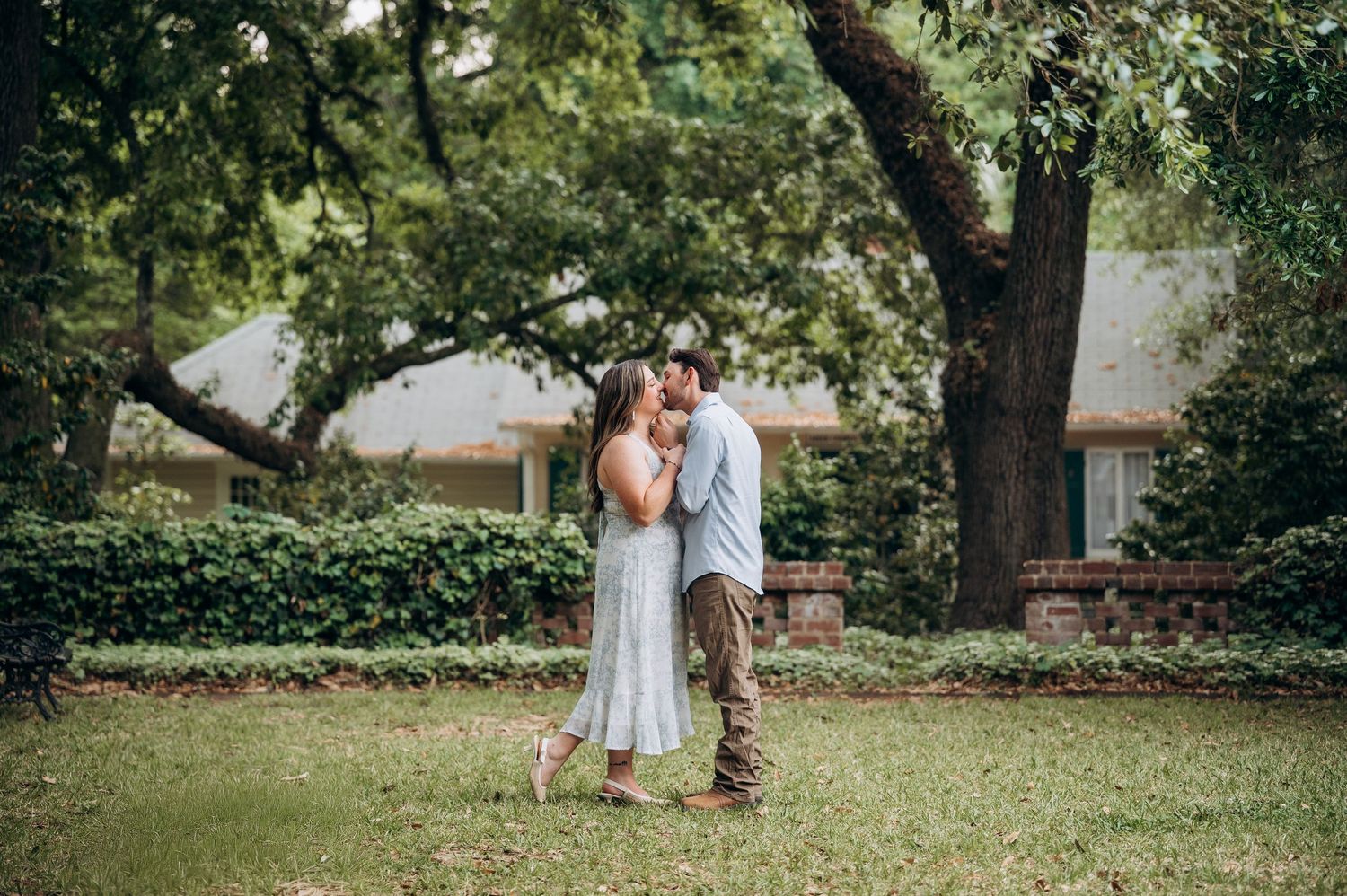 Couple sharing a kiss on a grassy lawn surrounded by green trees and shrubs.