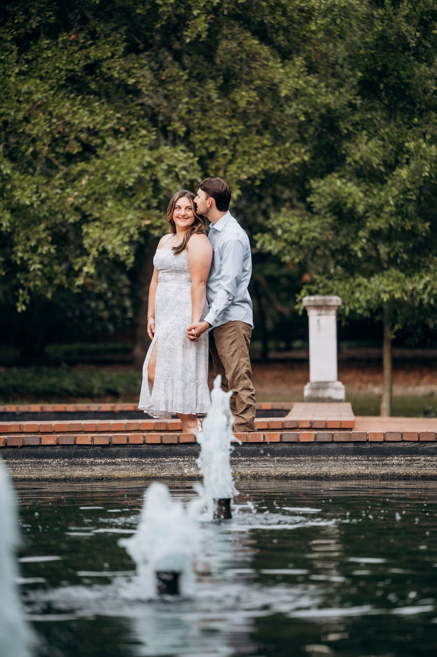 Romantic couple holding hands near a garden fountain, man kissing woman's cheek outdoors.