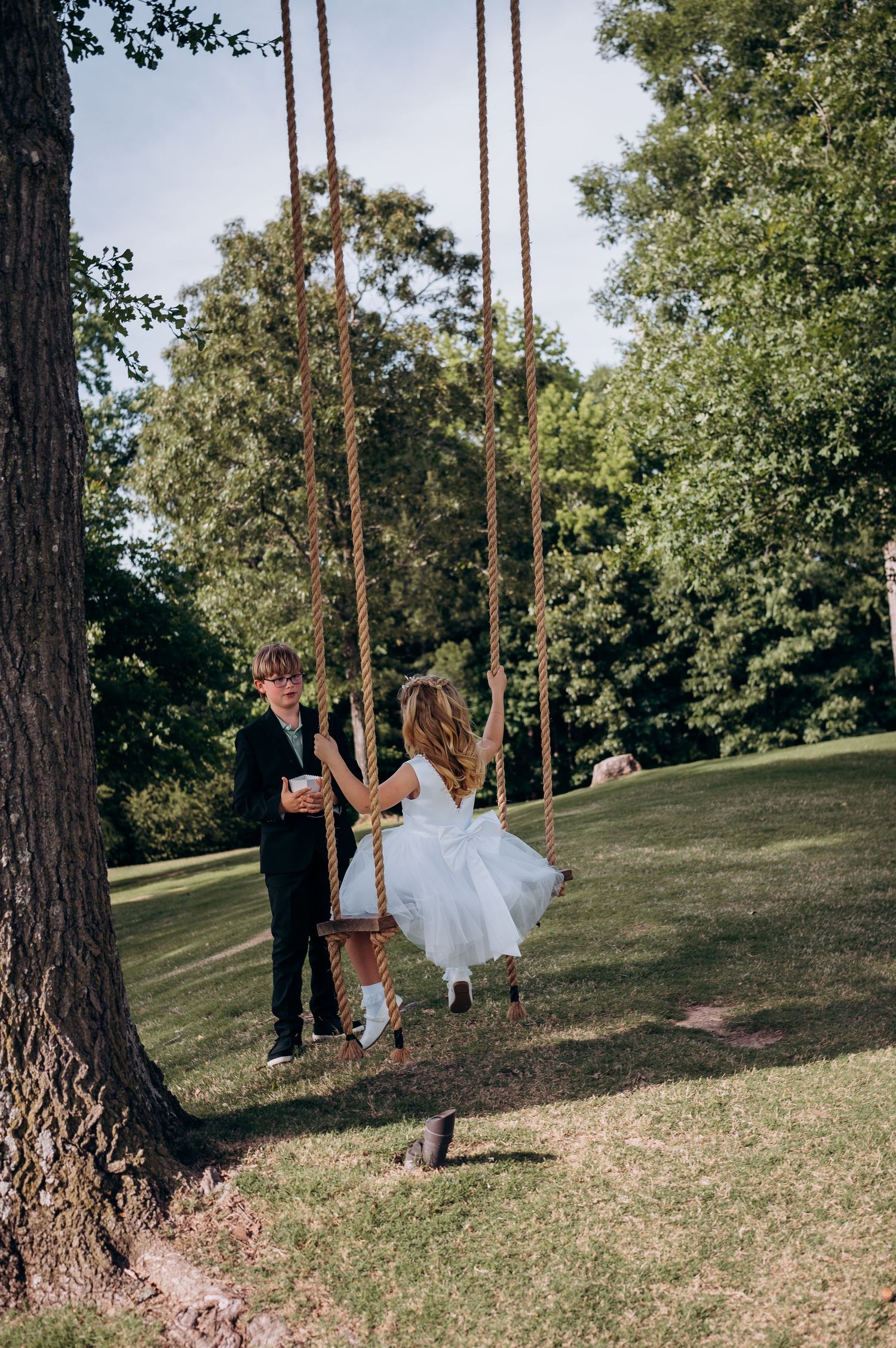 A bride in a white dress swings on a rope swing while a groom in a black suit stands beside her in a lush garden.