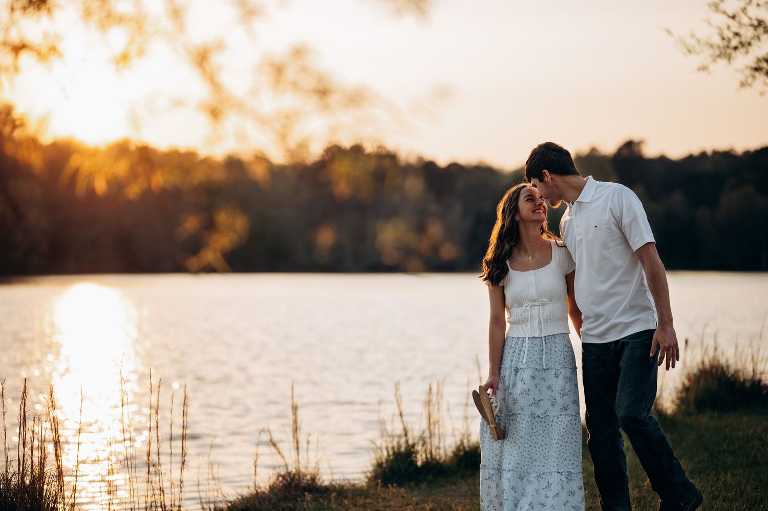 Couple embracing at golden hour sunset by a serene lake, woman in white floral dress, man in white shirt.