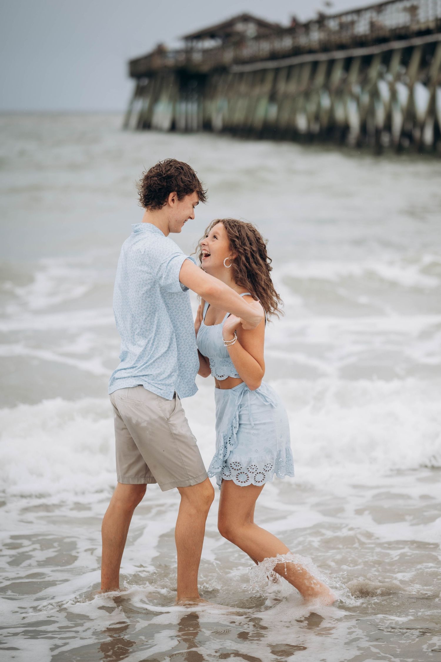 Couple splashing and playing in the ocean waves near a pier on an overcast day.