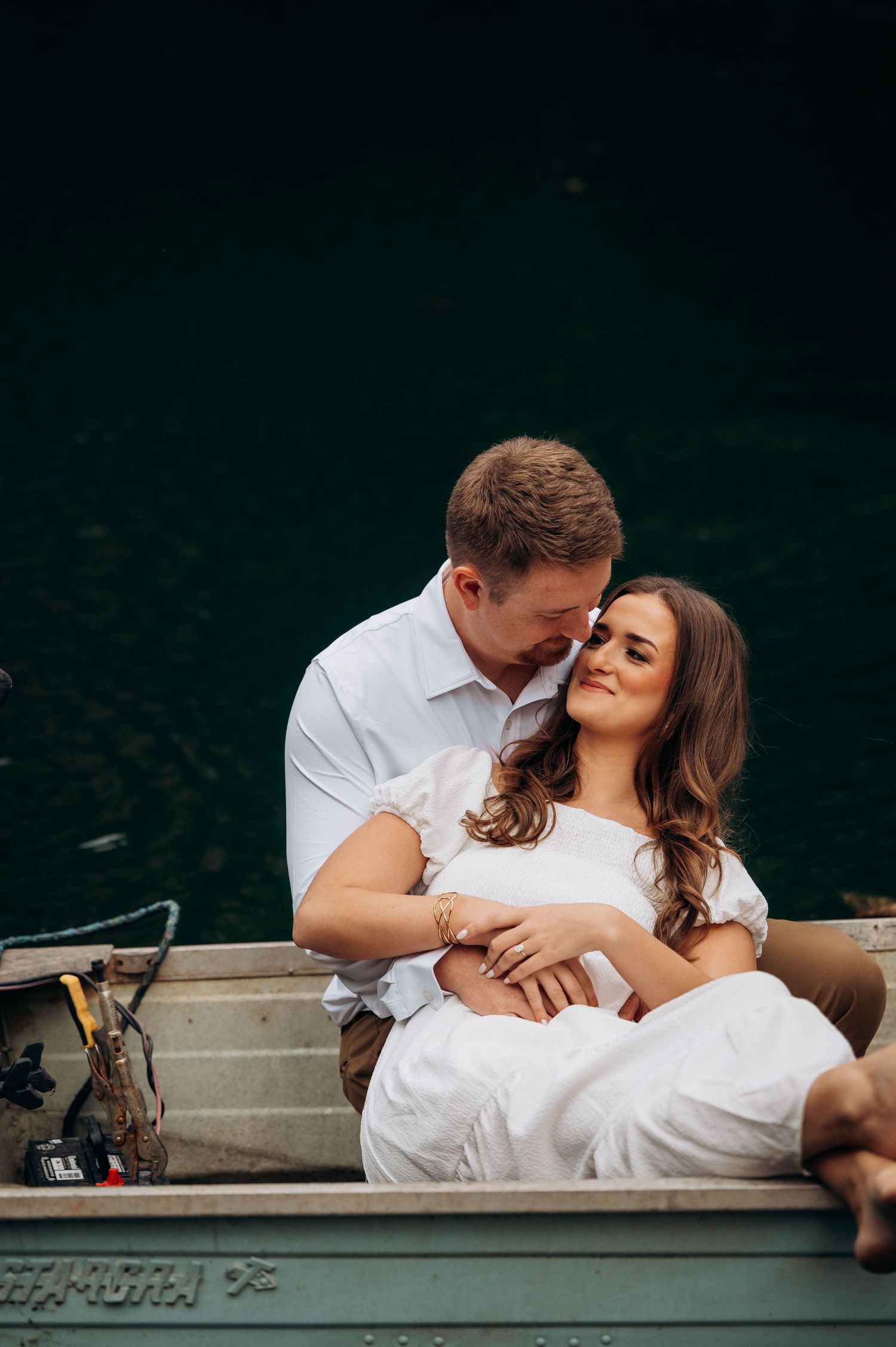 Couple in white clothing cuddles together in a small boat on dark waters.