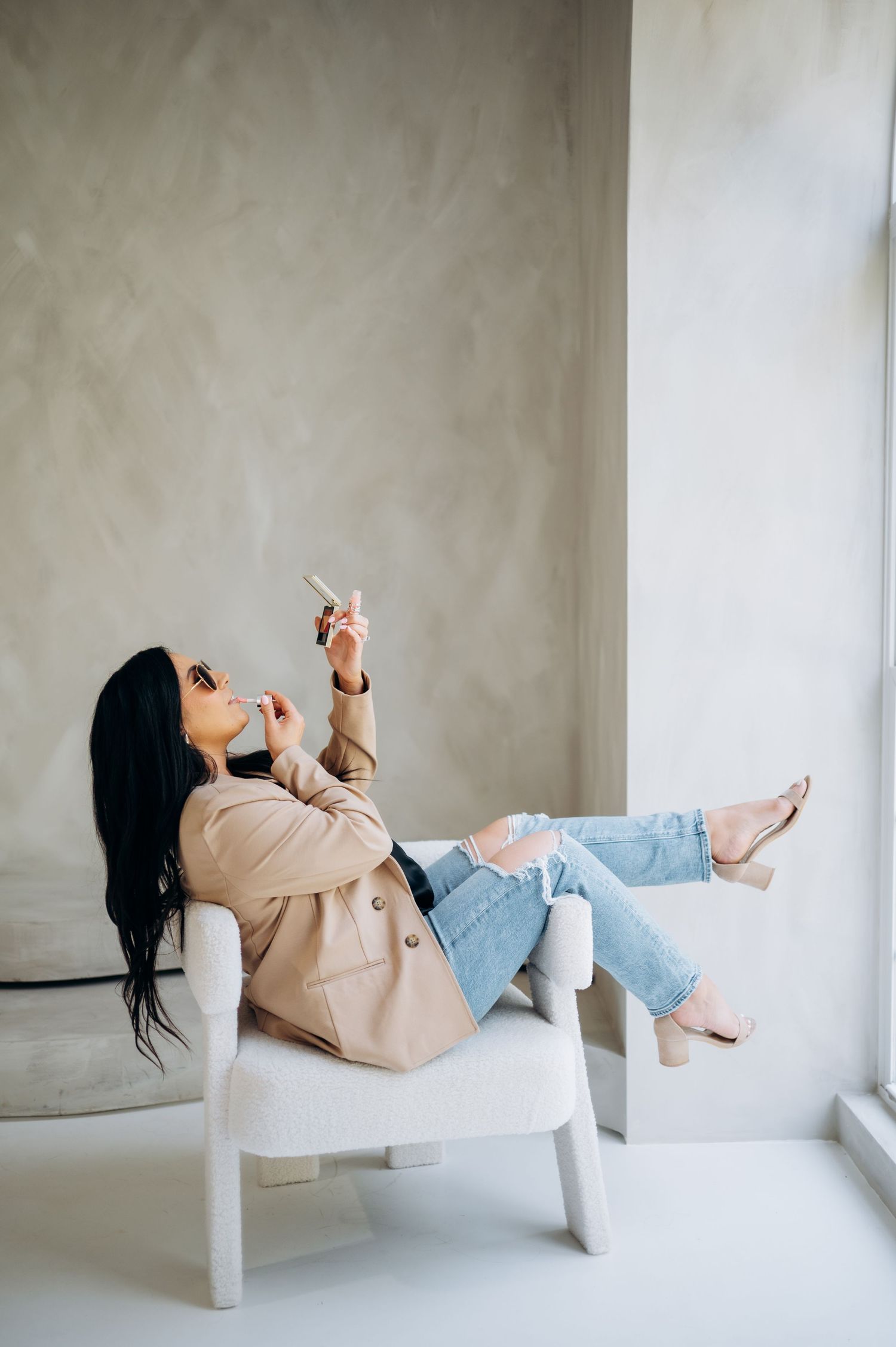 Relaxed pose in beige blazer and ripped jeans while sitting in white chair by window.