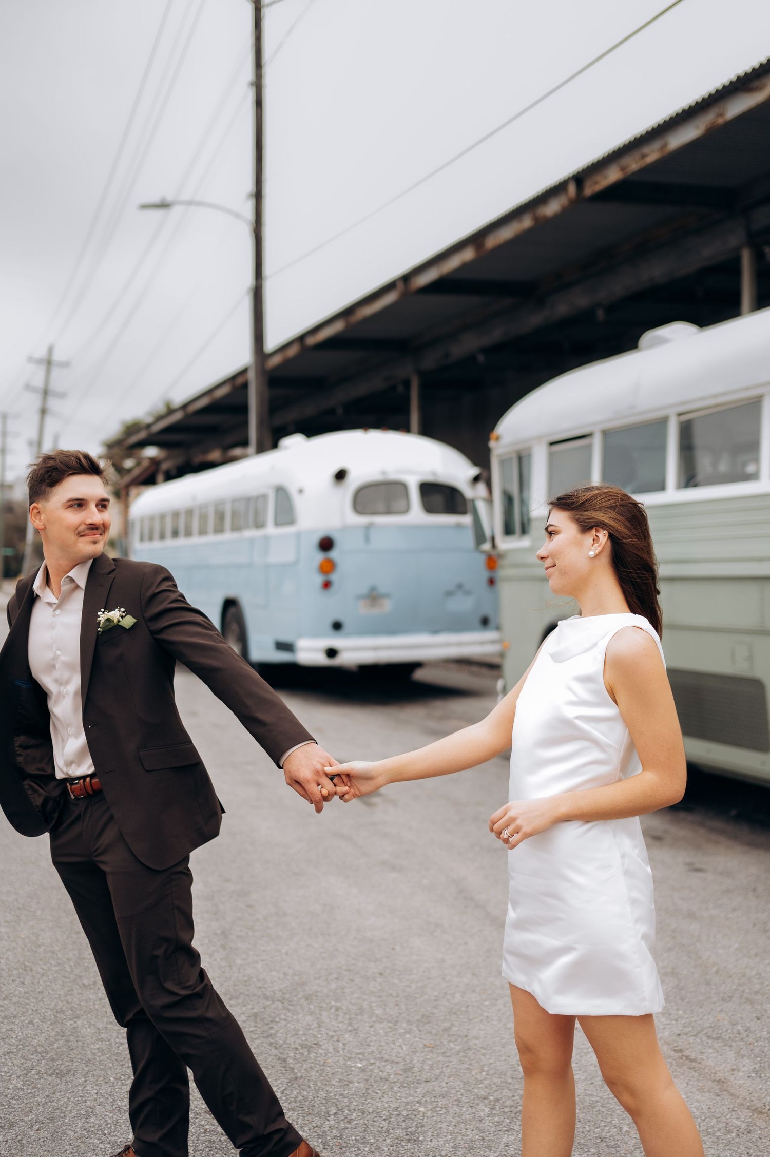 Playful couple in formal wear dance and spin in an industrial setting with vintage buses in background.