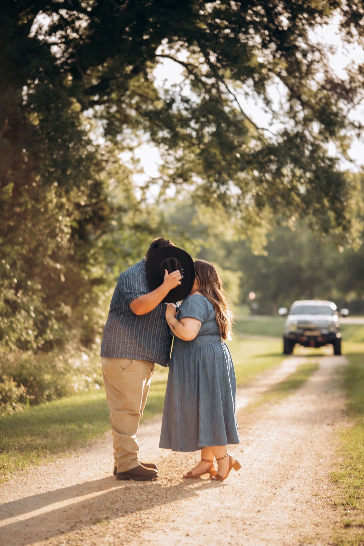 Couple sharing a kiss on a sunlit gravel path surrounded by lush green trees during golden hour.
