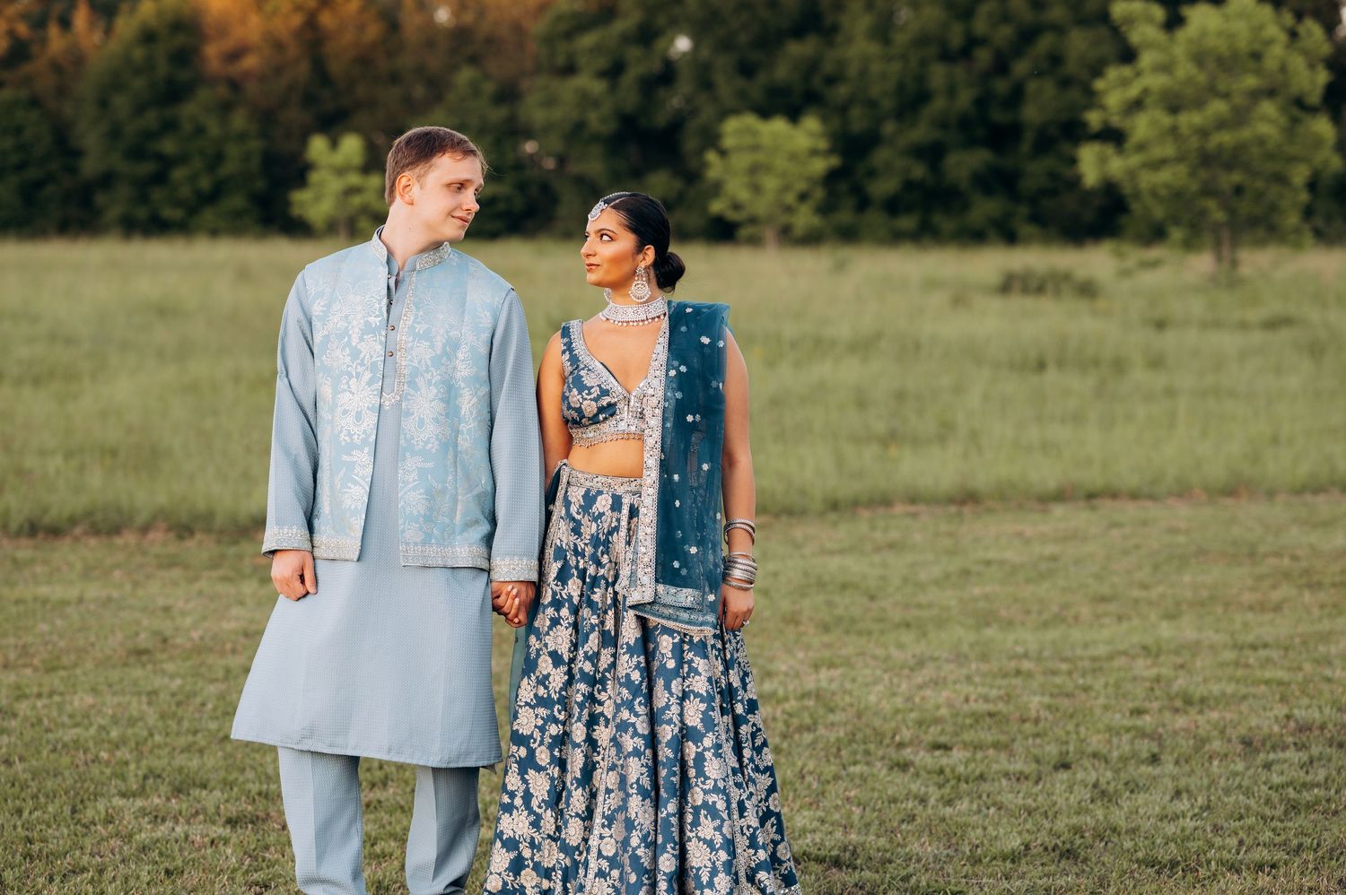 Couple in Indian wedding attire standing in green field, man in light blue kurta, woman in blue floral lehenga.