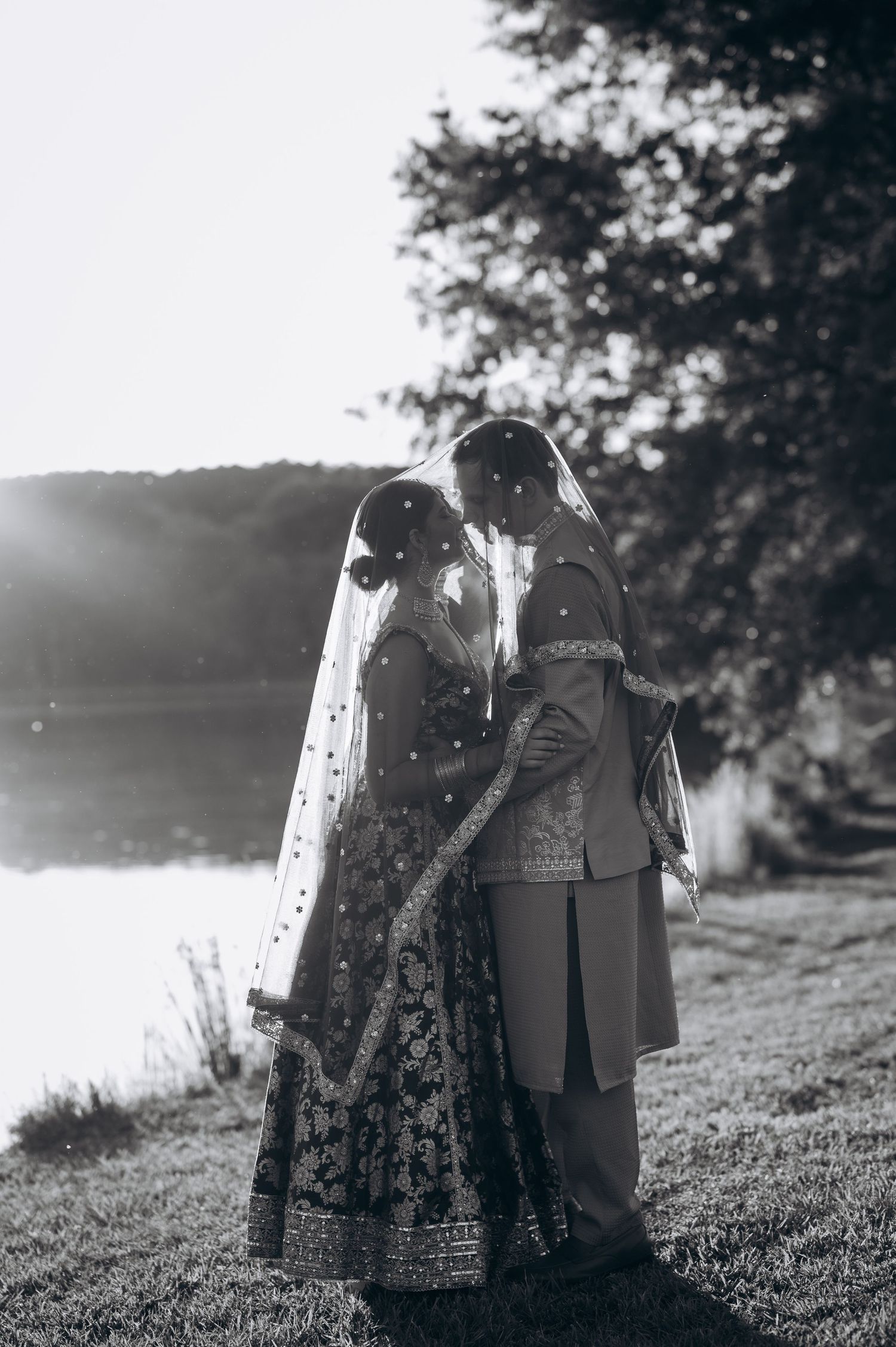 Black and white photo of South Asian bride and groom sharing an intimate moment under veil by a lakeside.