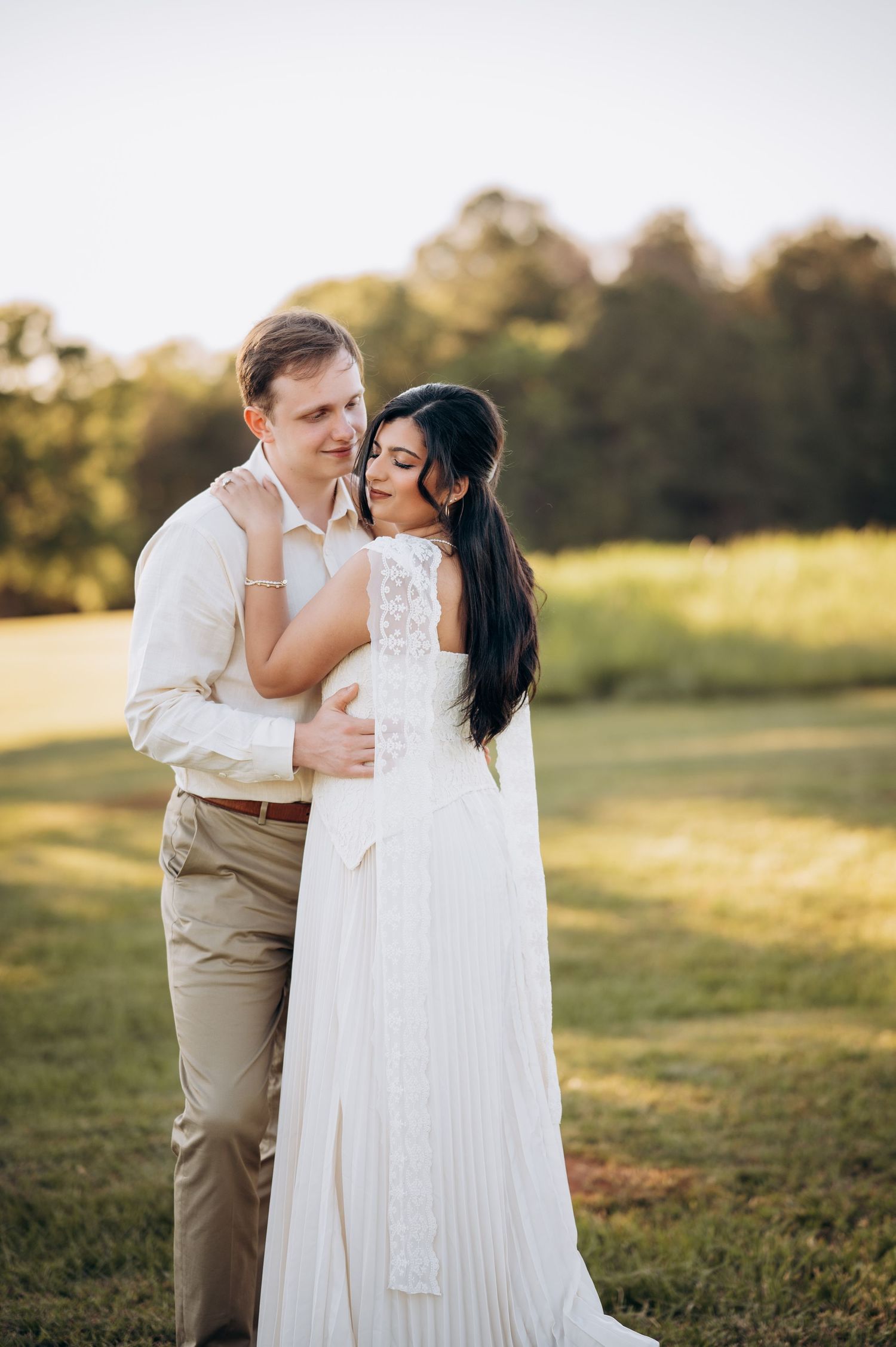 Couple shares romantic embrace in sunlit field, bride in flowing white lace dress, groom in tan suit.