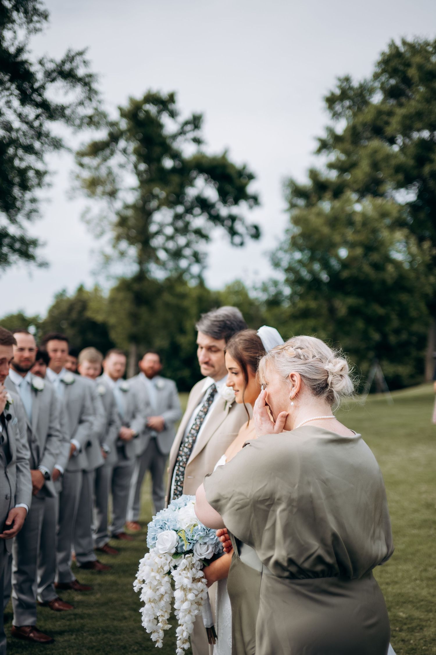 Bride holding blue hydrangea bouquet with wedding party during outdoor ceremony.