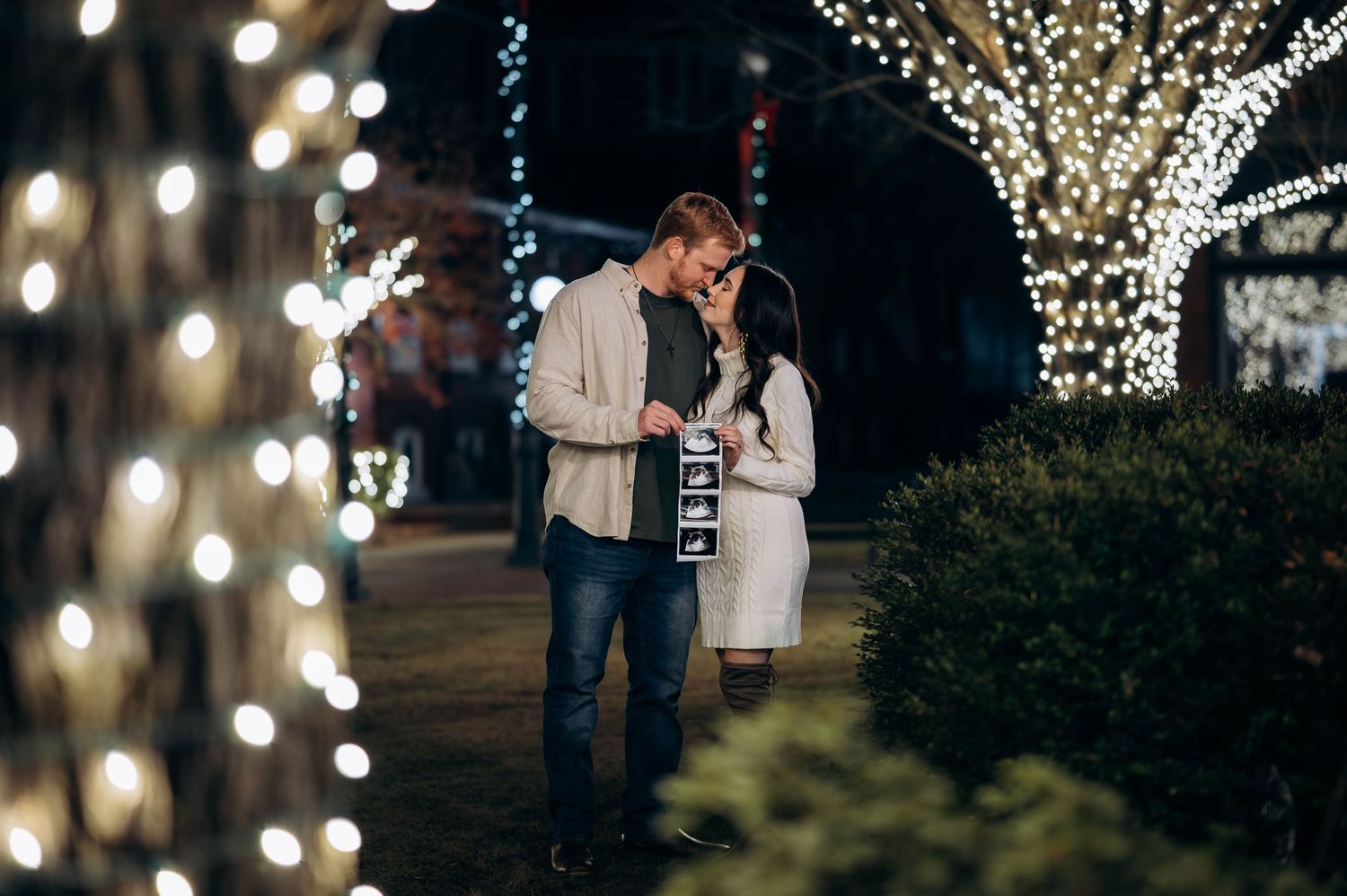 Couple sharing a romantic moment under glowing Christmas lights, holding ultrasound photos.