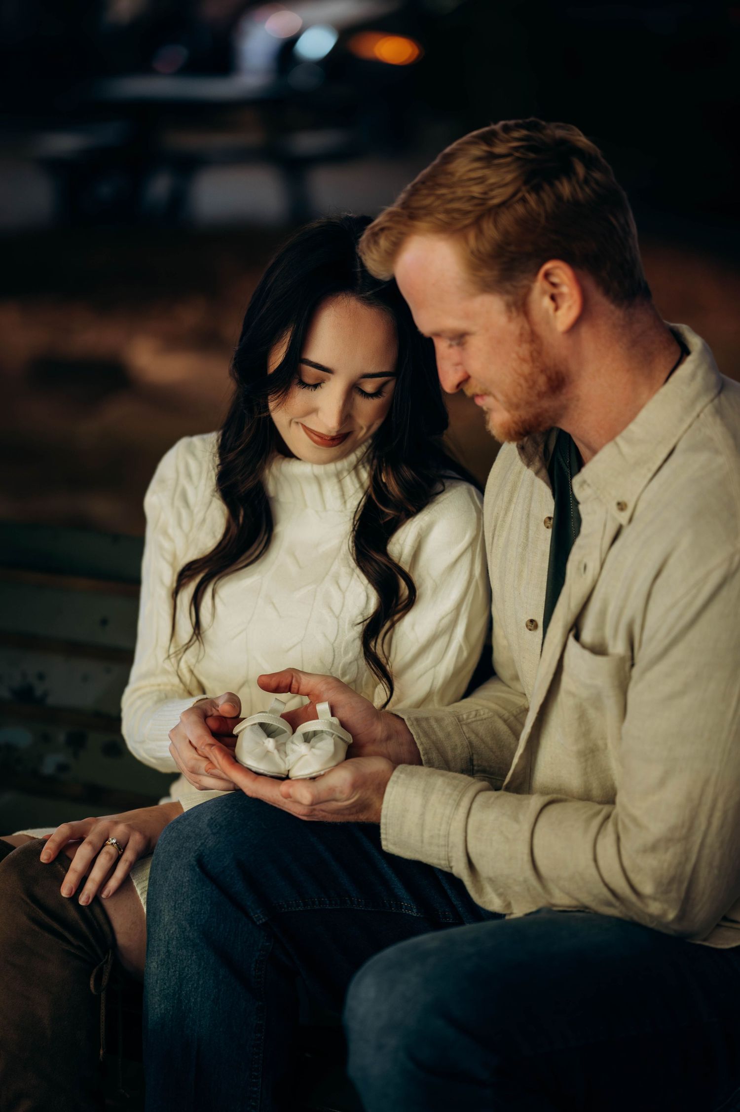 A couple sits together at night, tenderly holding tiny baby shoes, announcing their pregnancy in a romantic outdoor setting.