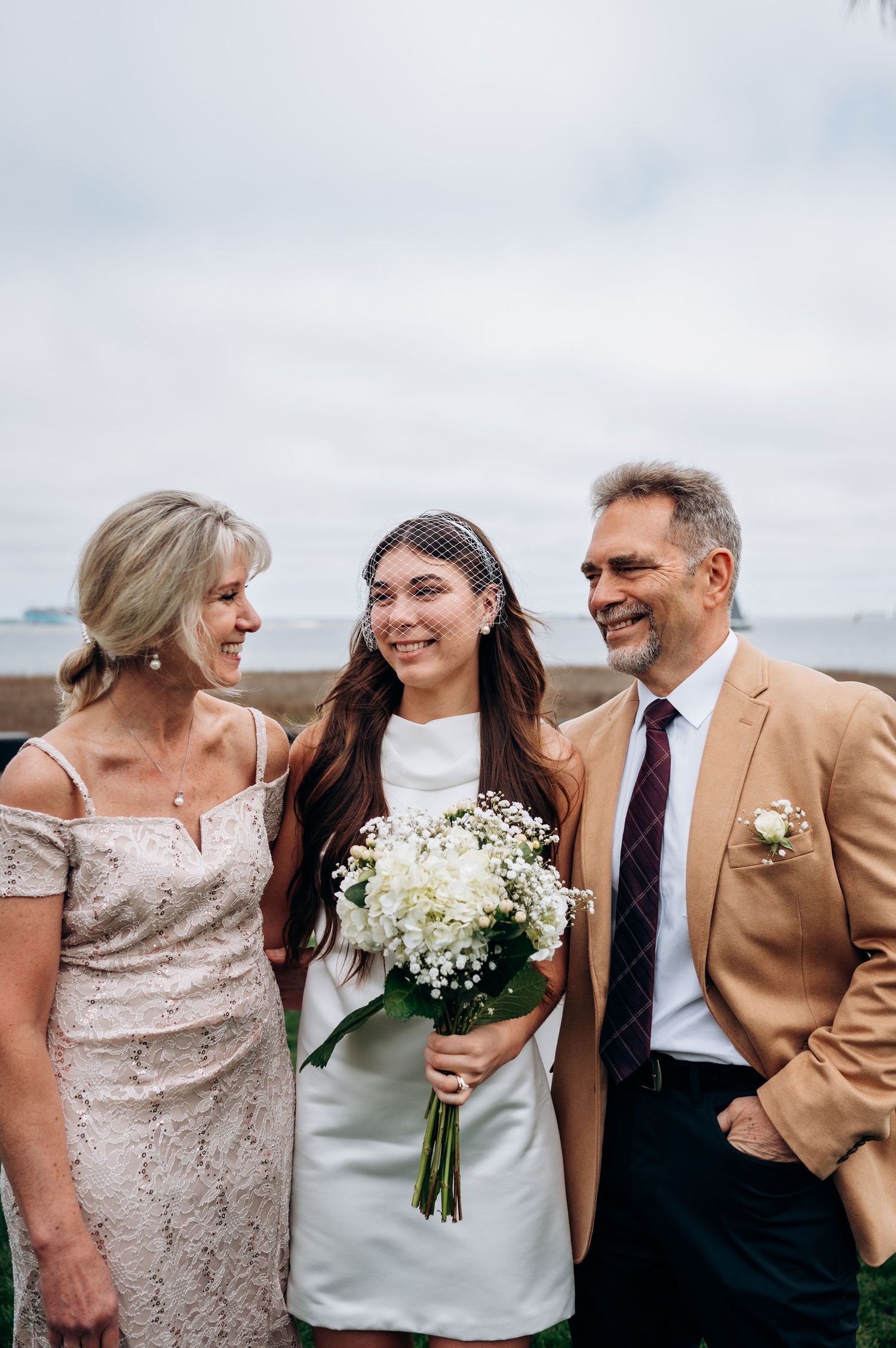 Happy bride in white mini dress holds white bouquet flanked by smiling parents at outdoor waterfront wedding.