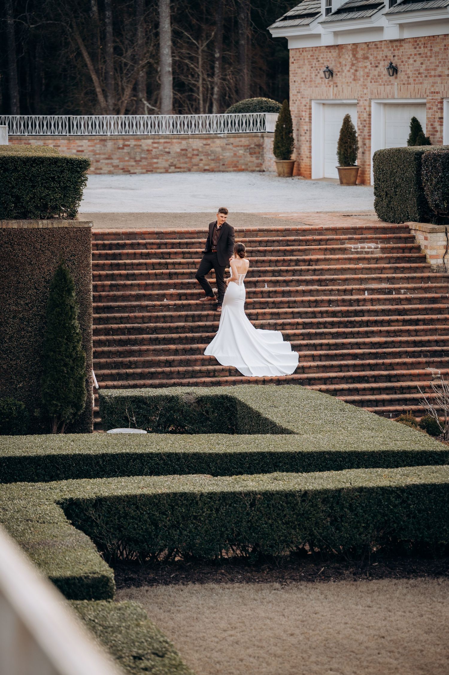 Bride in flowing white gown and groom ascending grand brick staircase in formal garden.