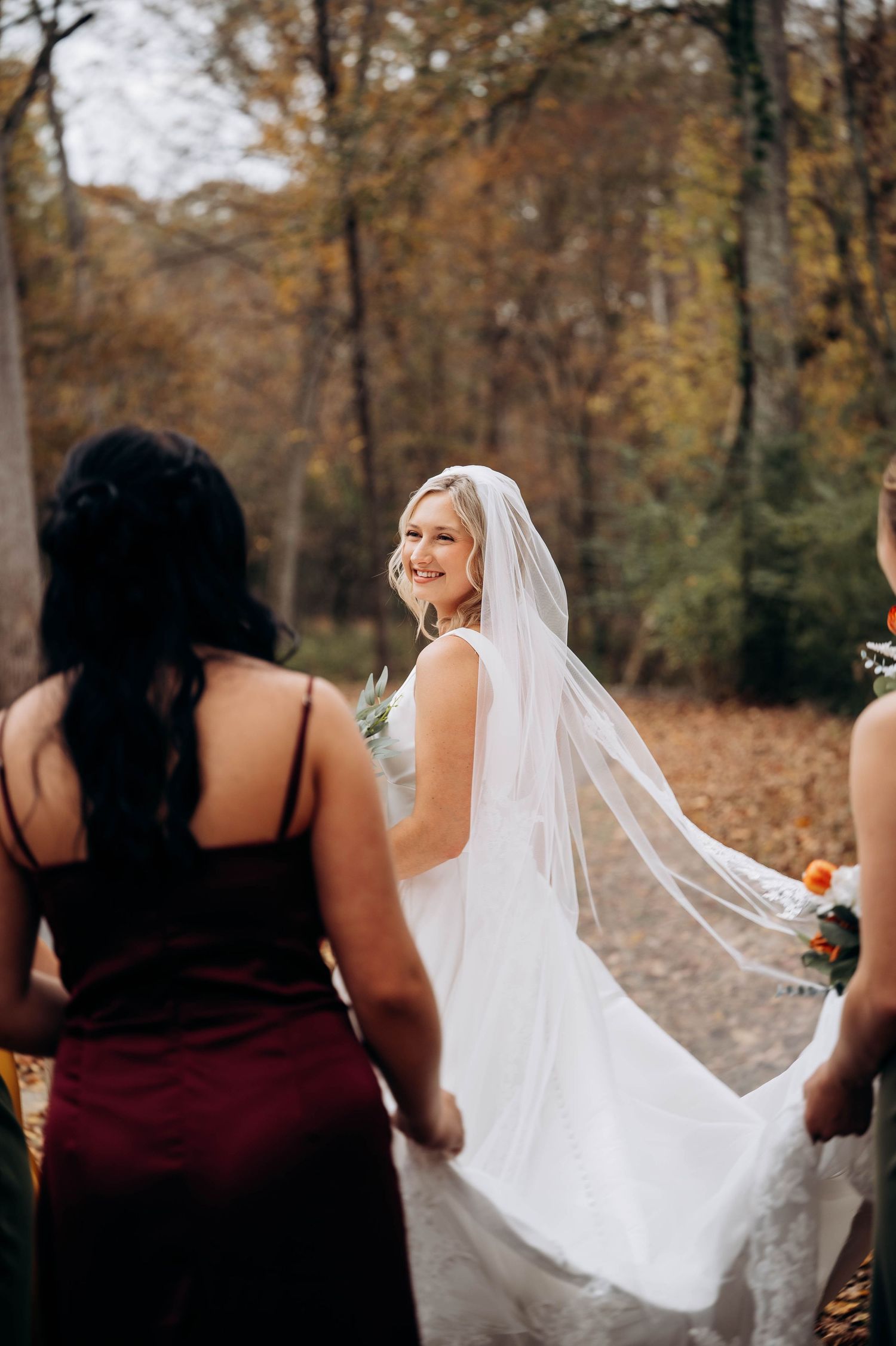 Bride in white dress with flowing veil surrounded by bridesmaids in burgundy dresses during fall photoshoot.