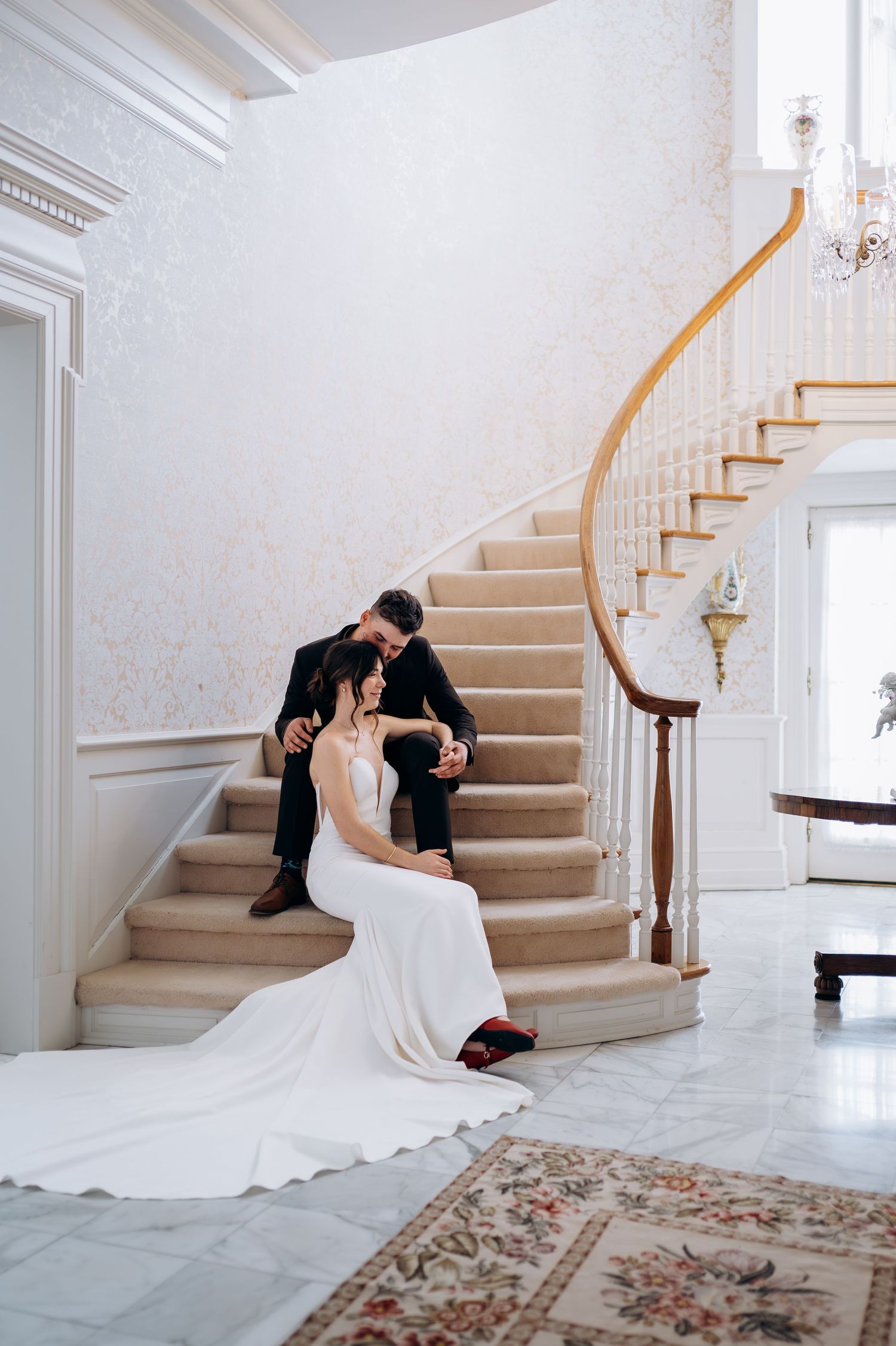 Bride and groom sitting together on elegant curved staircase in white wedding dress and black suit.