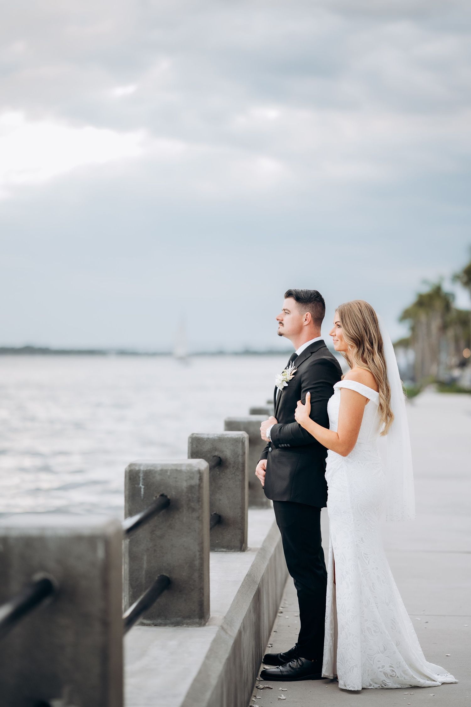 Bride and groom stand together on a waterfront promenade, gazing at the ocean under a cloudy sky.