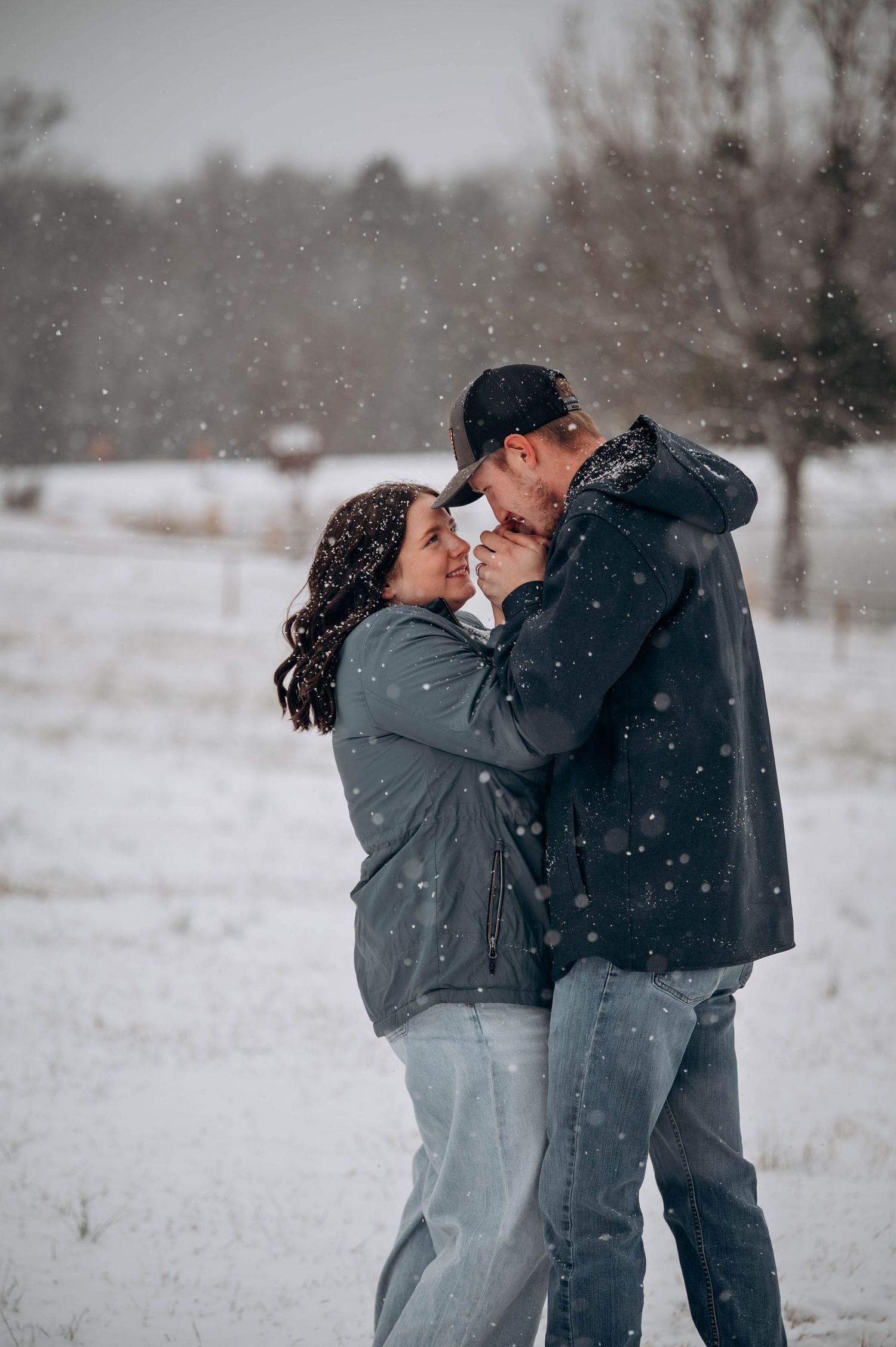 A couple shares a loving embrace while snowflakes fall around them in a peaceful winter setting.