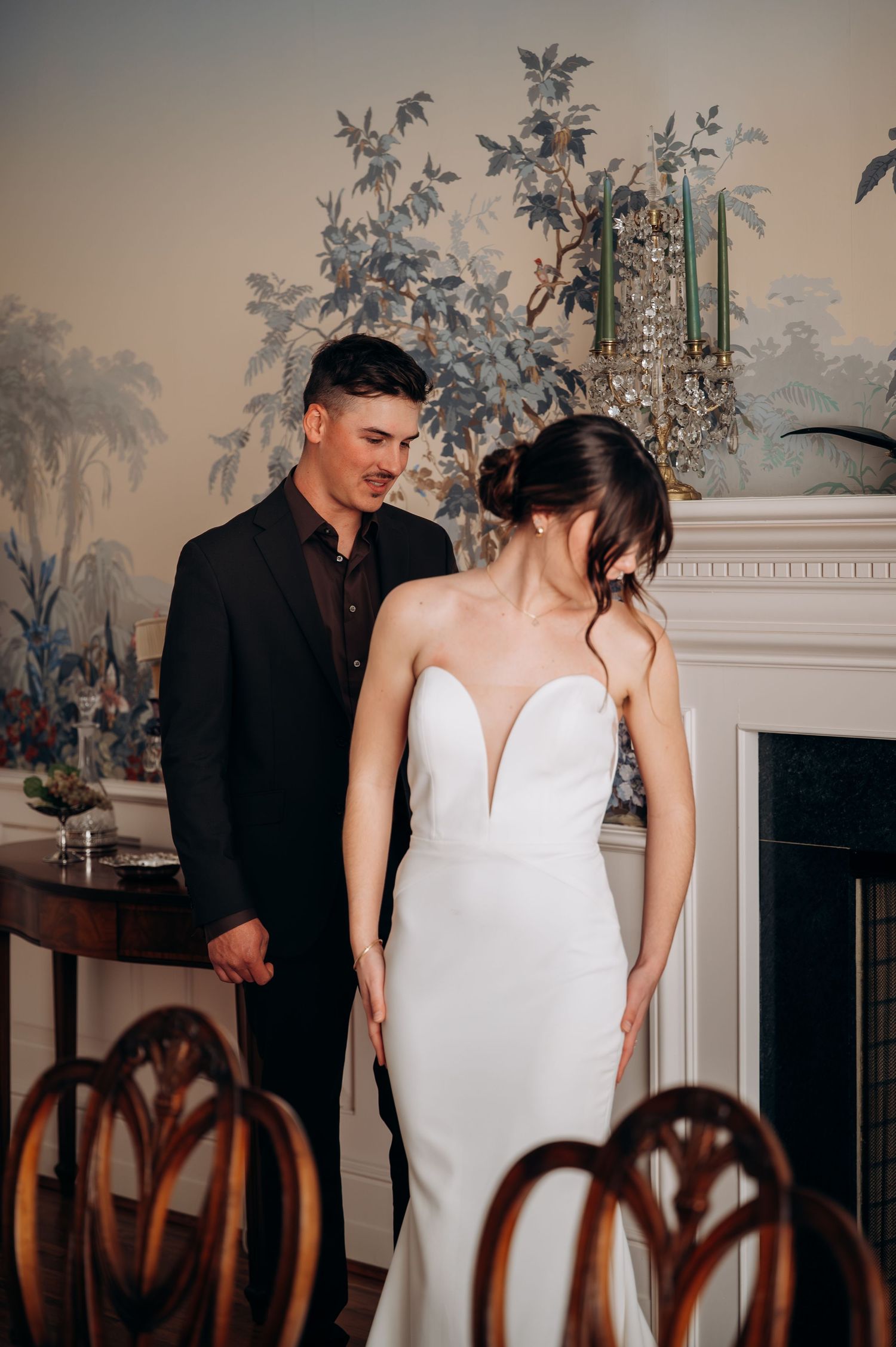 Elegant couple in formal attire poses in front of a classic fireplace with vintage decor.