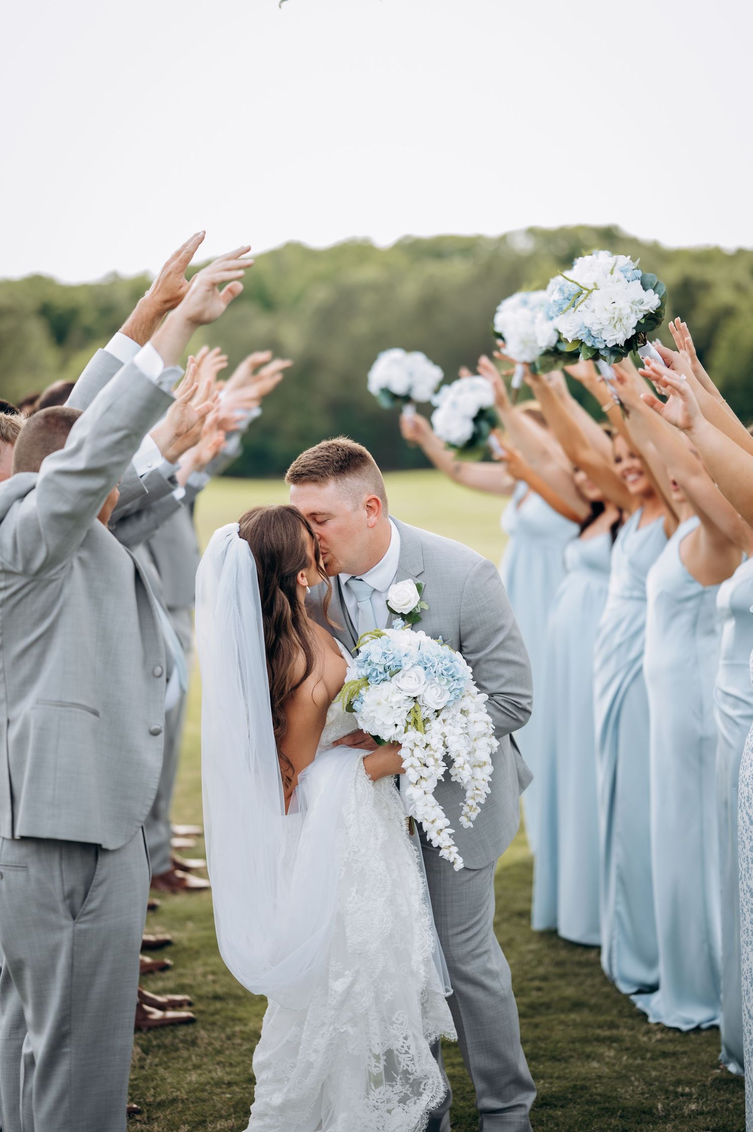 Bride and groom share a kiss while wedding party in gray suits and blue dresses raise bouquets in celebration outdoors.