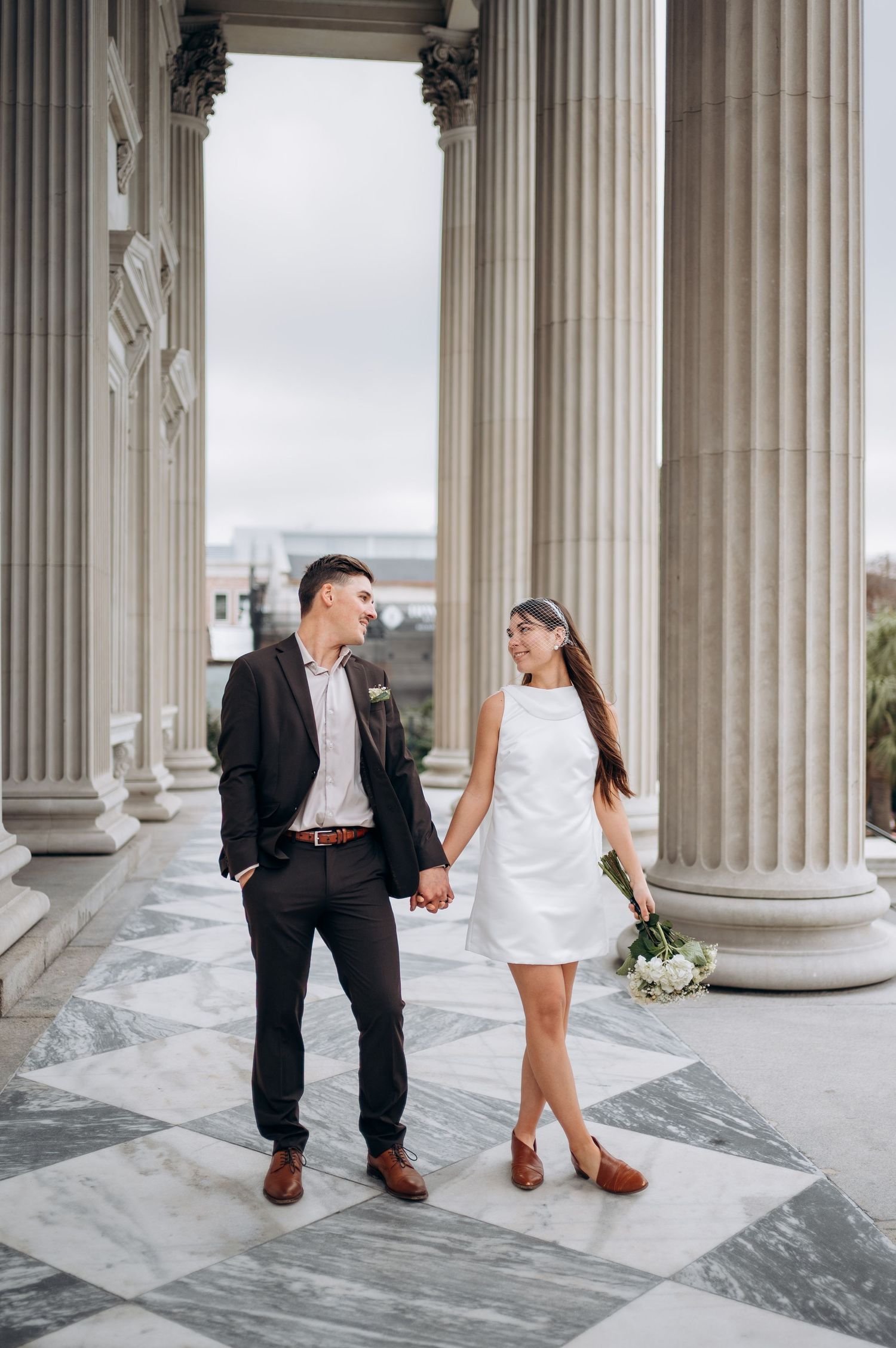A newlywed couple poses between classical columns in formal wedding attire.
