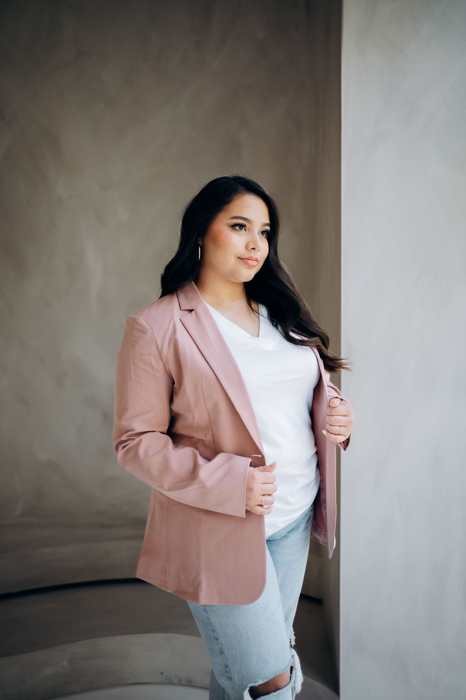 Professional portrait in a pink blazer and white top against a textured gray wall, styled with distressed jeans.