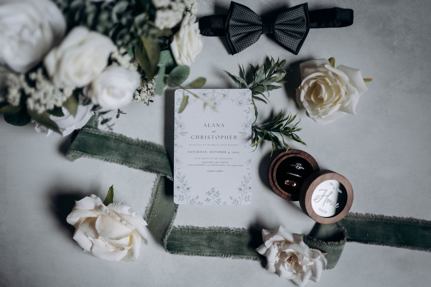 Wedding flatlay with black bowtie, white roses, and invitation on velvet fabric.