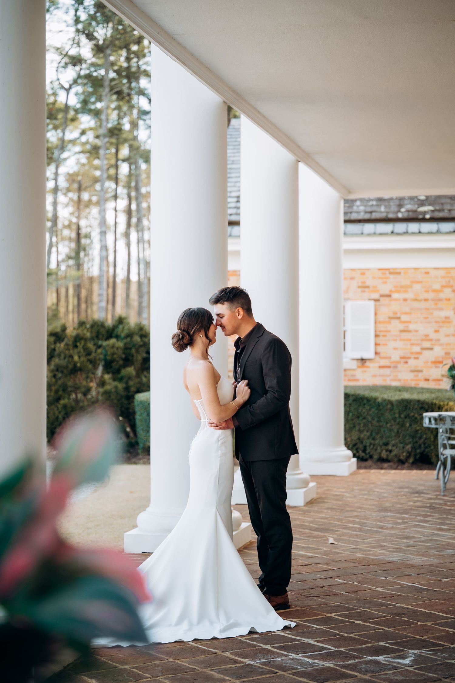 Wedding couple embraces on a columned porch with brick details and pine trees in background.