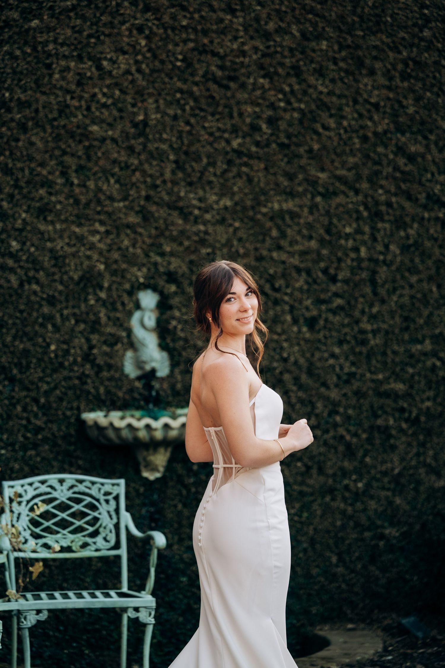 Bride in white dress poses near vintage mint green garden chairs against dark hedge backdrop.