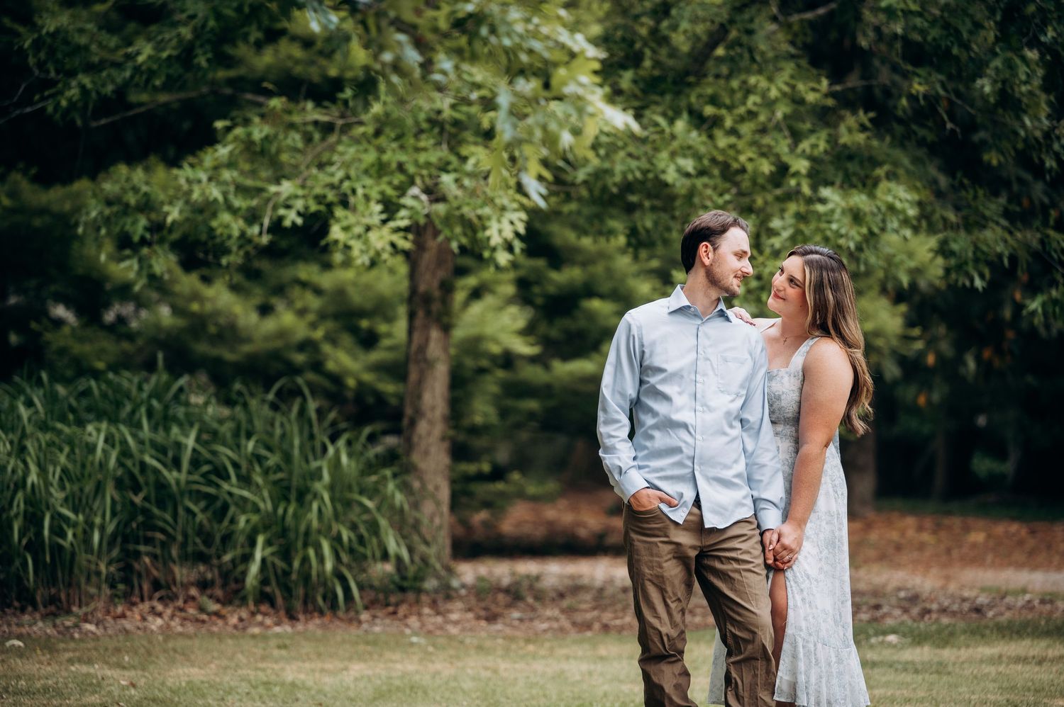 Couple laughing and gazing at each other while standing on a grassy lawn with tall green trees.