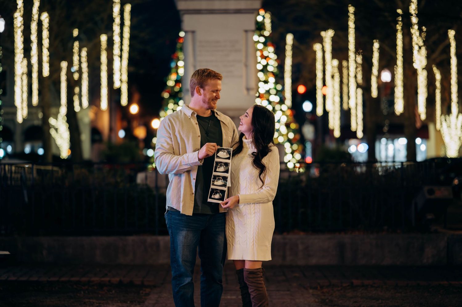 Couple sharing a joyful moment holding ultrasound photos amid festive holiday lights at night.