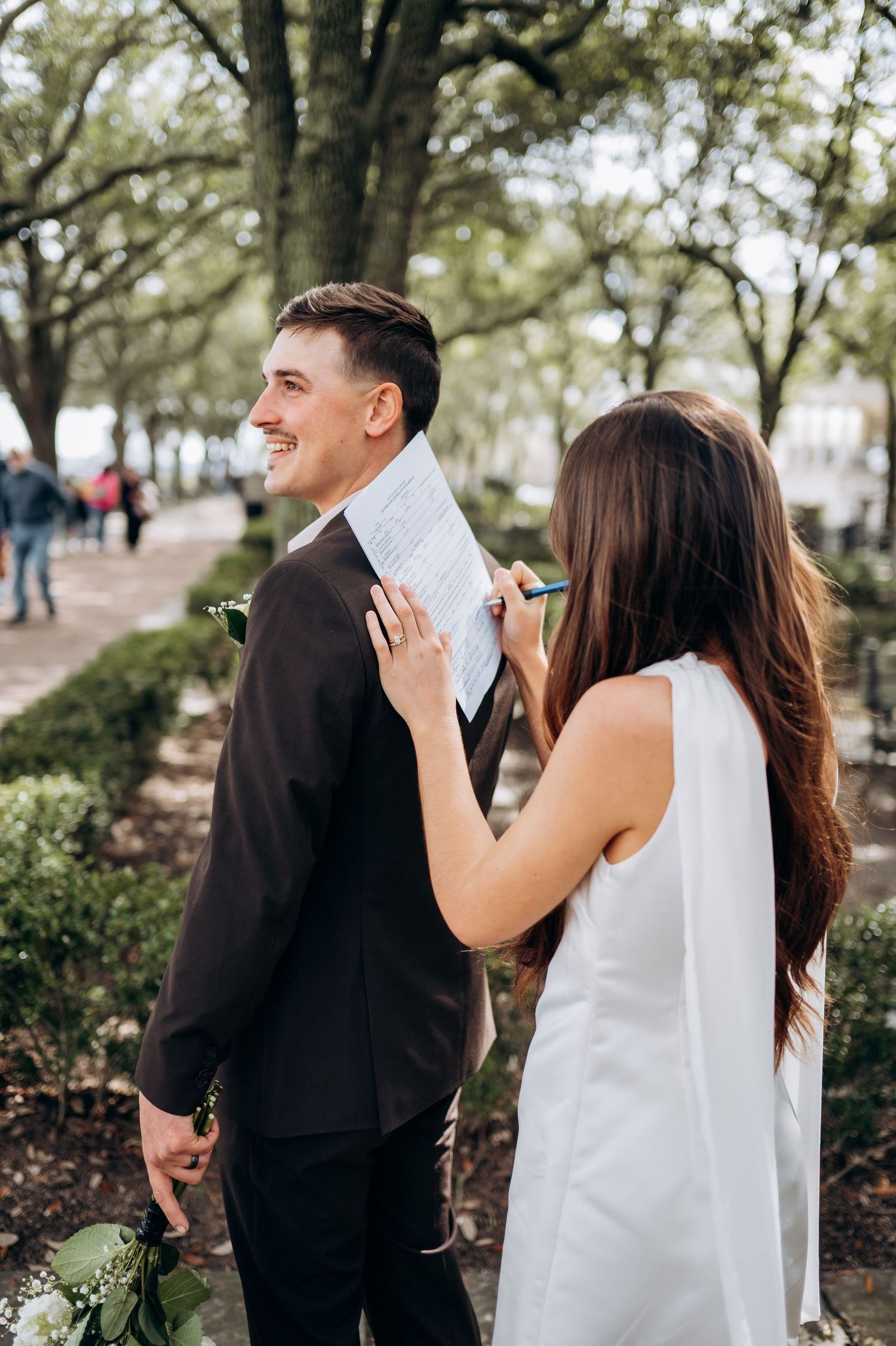 Bride signs marriage license on groom's back in a park, both smiling, wearing wedding attire outdoors.