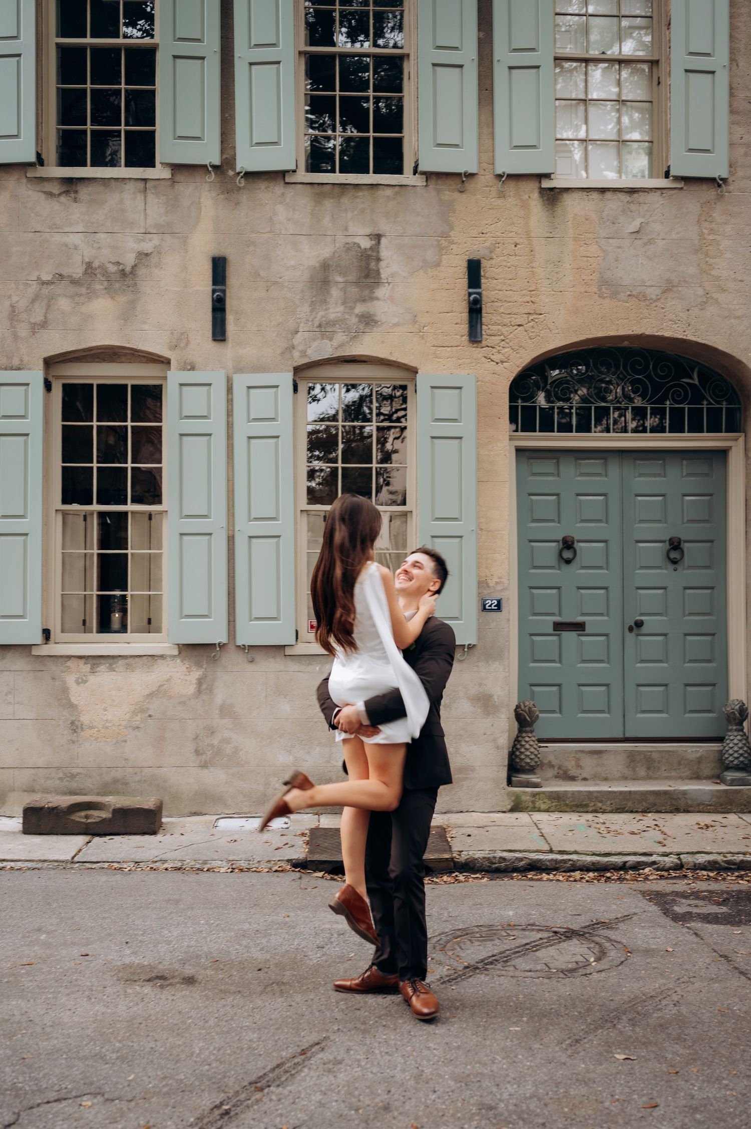 Groom lifts smiling bride in white dress outside a charming building with sage green shutters and door.