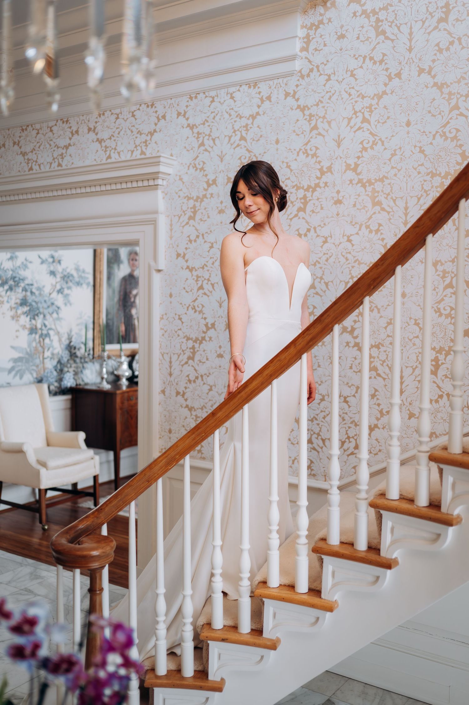 Bride in sleek white strapless gown with plunging neckline poses gracefully on elegant staircase with chandelier.