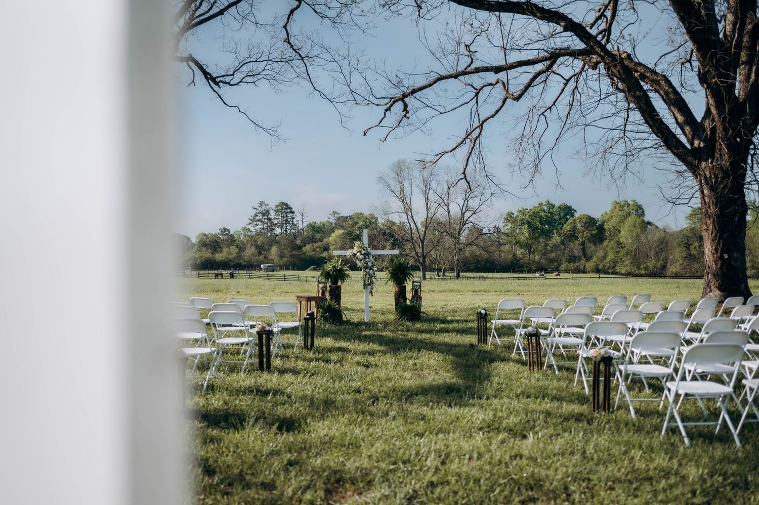 Outdoor wedding ceremony setup with white folding chairs on green grass near a large tree overlooking a serene pond.