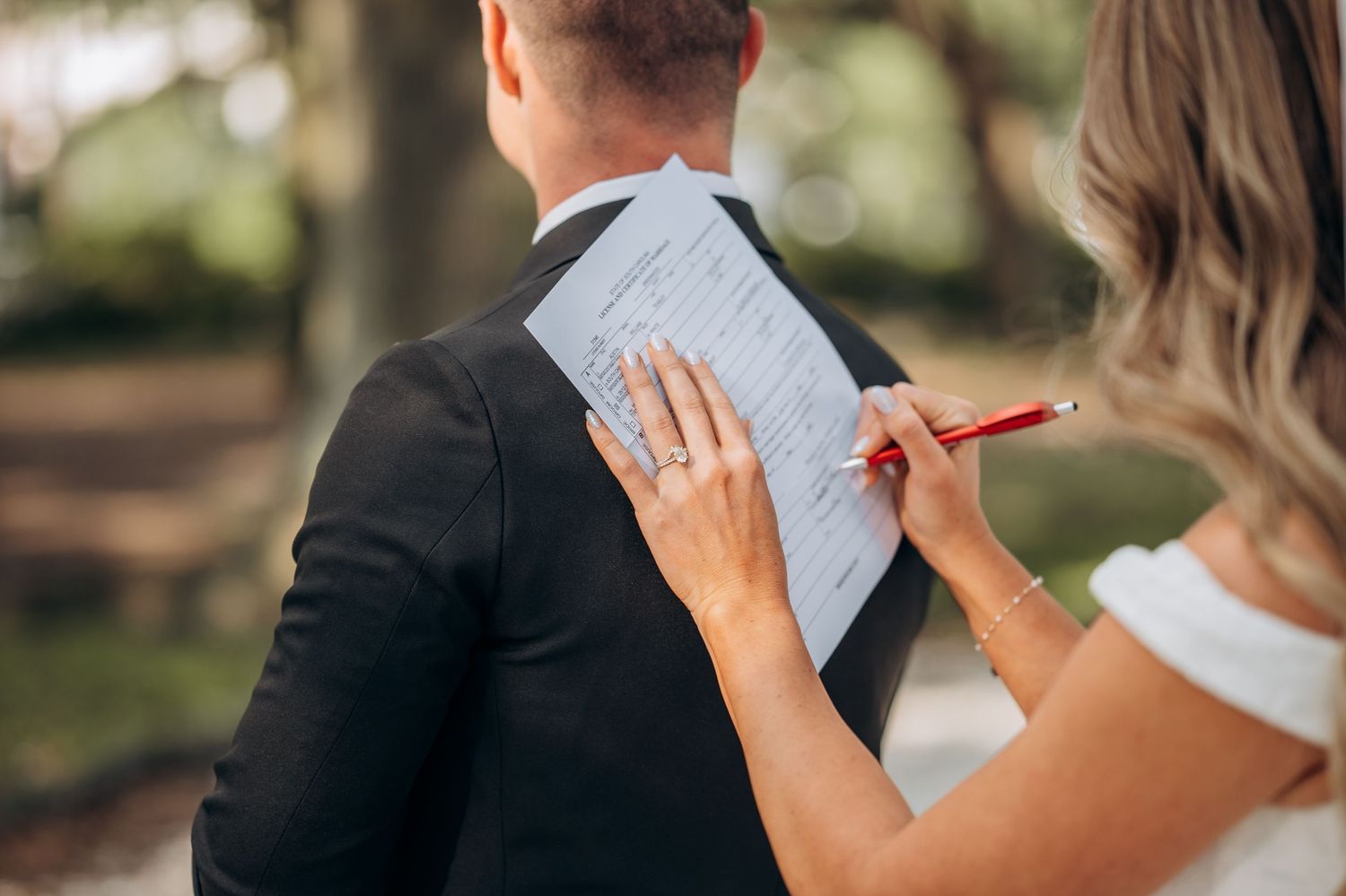 Bride signing marriage certificate on groom's back outdoors, wearing engagement ring, holding red pen.